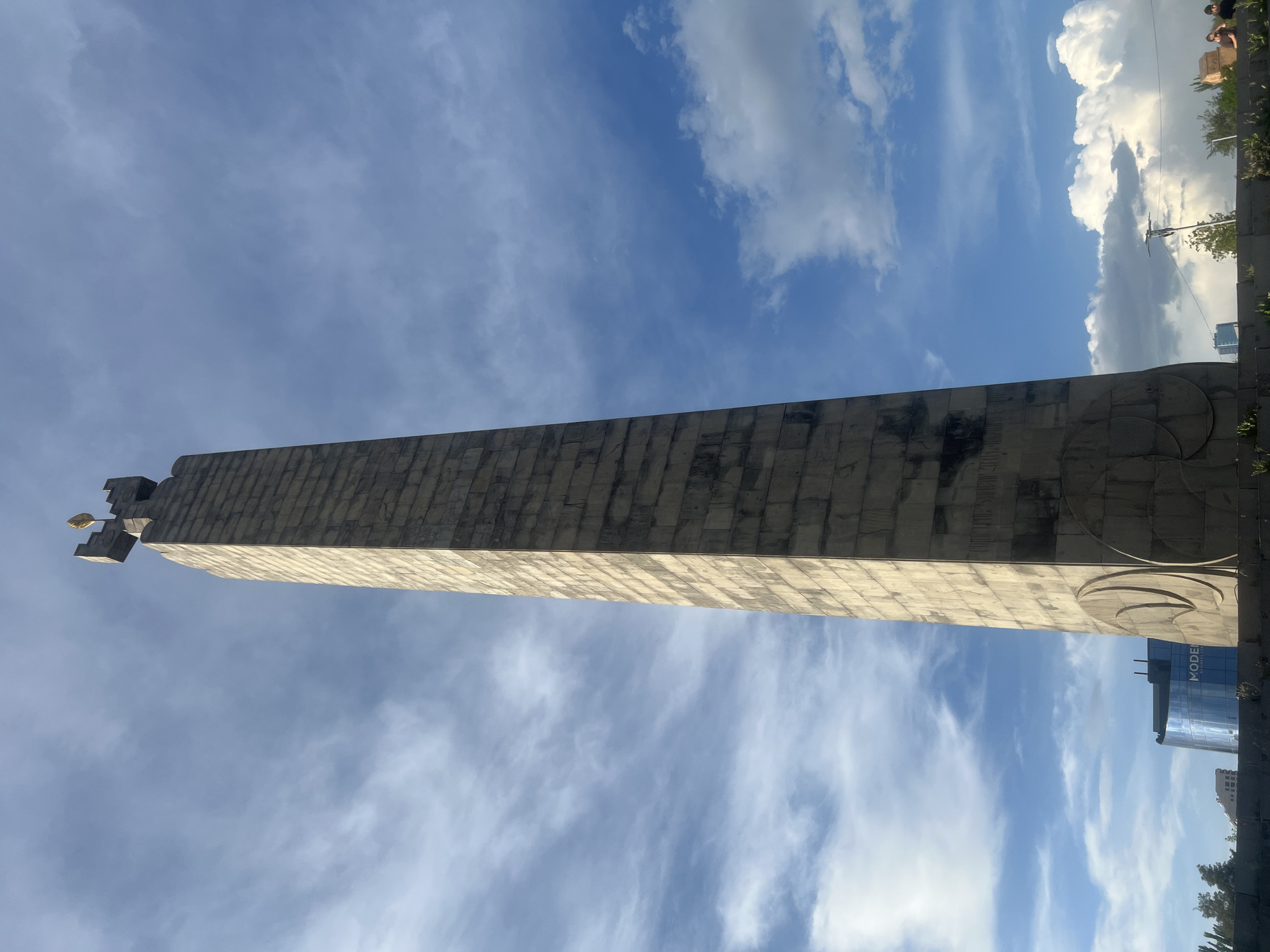 The image depicts the Victory Monument, a prominent obelisk located in Haghtanak Park (Victory Park) in Yerevan, Armenia. The monument is a tall, slender, rectangular obelisk constructed from light-colored stone blocks, exhibiting signs of weathering and discoloration. Its base features large, stylized circular relief carvings. The apex of the monument is topped with a smaller, crenellated structure, reminiscent of castle battlements, from which a golden, flame-shaped or leaf-shaped ornament protrudes vertically.
The monument stands against a dynamic sky, characterized by a mix of light blue areas and scattered white clouds. Towards the upper left, the clouds are wispy and diffuse, while towards the bottom right, they become thicker and more defined cumulus formations.
In the mid-ground, to the left of the monument, a modern building with a curved, blue glass facade is visible, bearing the partial inscription "MODERN BUSINESS CEN" (likely "MODERN BUSINESS CENTER"). Further left, dark green foliage from trees can be seen. To the right, indistinct dark trees and some brownish structures are present in the distance. The foreground shows a low, stone wall or elevated platform, partially covered with sparse green vegetation. Near the bottom center, a thin pole with an antenna-like apparatus extends upwards. On the far right at ground level, several small, indistinct human figures are visible, suggesting people are in the vicinity, but their specific actions or interactions are not discernible. The scene is illuminated by natural daylight.