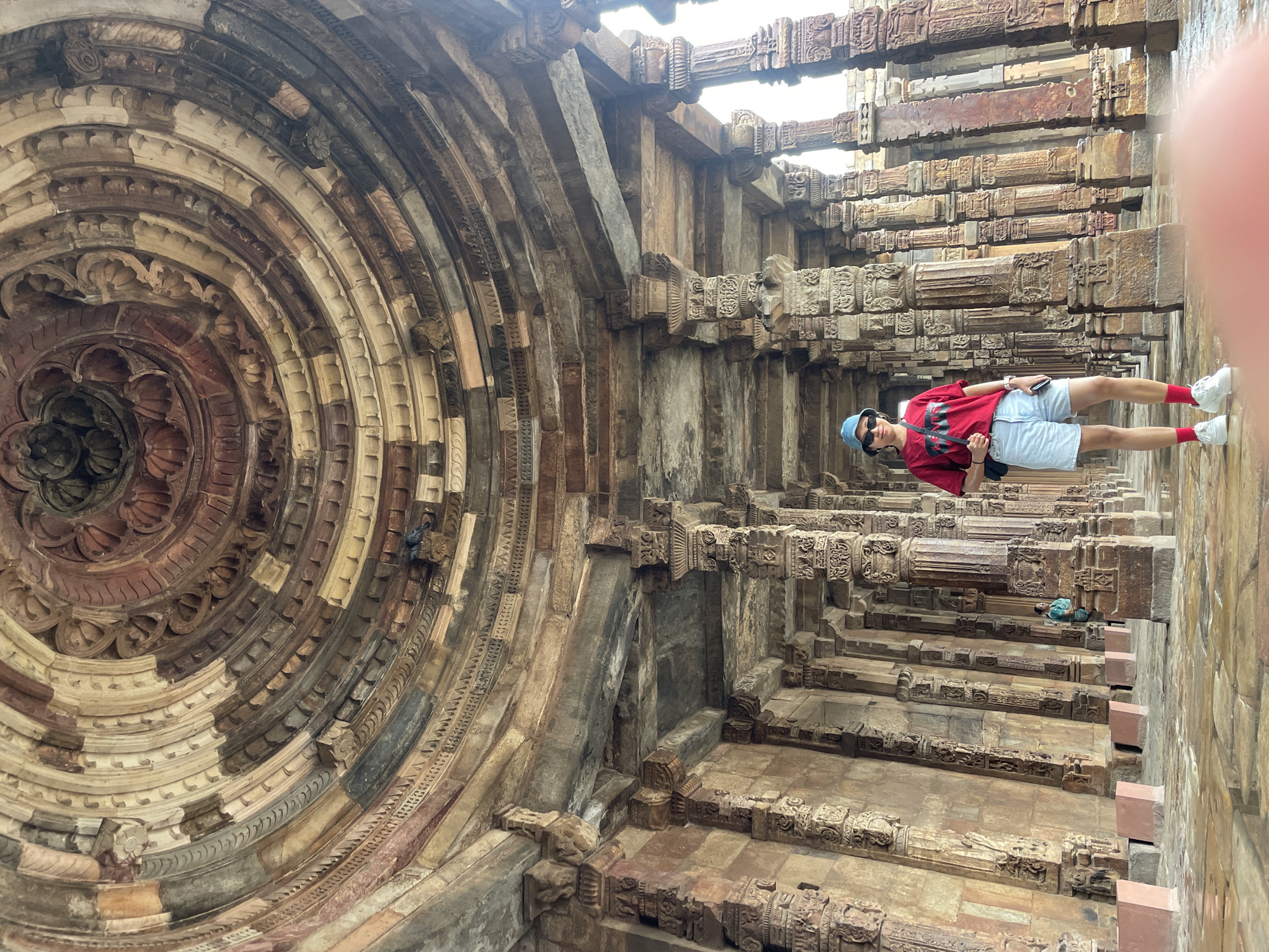 A full-shot, eye-level, vertical photograph captures a woman standing in the interior of the Qutub Minar complex in Delhi, India. The woman, positioned slightly left of center, wears a red t-shirt, light blue denim shorts, a blue bucket hat, white sneakers, and red socks. She carries a black crossbody bag and holds a smartphone in her right hand. Her posture is relaxed, with her weight on her left leg and her right leg slightly bent.
The surrounding environment is characterized by ancient architecture, featuring intricately carved stone pillars that support an ornate, circular, domed ceiling. The pillars are arranged in a colonnade, receding into the background. The ceiling displays complex geometric and floral carvings in various shades of brown and beige. The floor is wet and reflective, suggesting recent rain or a cleaning. In the mid-ground, to the left of the woman and further back, a second individual, dressed in a blue-green traditional Indian garment, is visible, also facing the camera. The lighting is bright, with sunlight filtering into the space, creating highlights and shadows that emphasize the texture and depth of the stone structures.