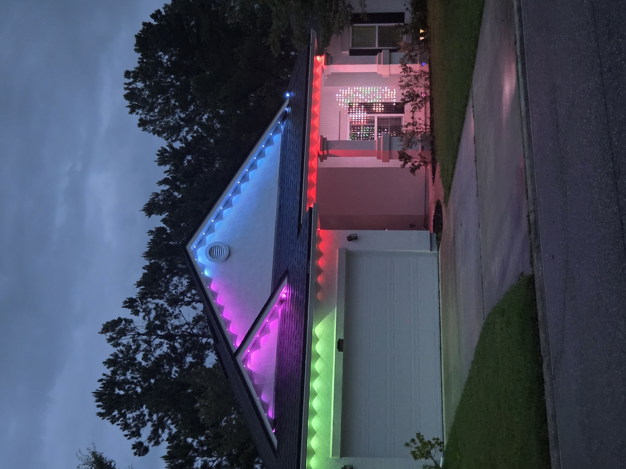 An exterior, eye-level, medium shot captures a white, two-story house adorned with Christmas lights in an unknown location. The sky above is overcast and dark blue, hinting at dusk or a stormy evening. Tall, dark green pine trees form a dense backdrop behind the house.
The house features a dark gray roof. The eaves are lined with colored LED lights: pink and blue on the upper sections and green on the lower, above the garage. The garage door is white and closed. A covered porch supported by two white columns is illuminated with red lights. Inside the porch window, a string of colorful Christmas lights forms a grid pattern, with individual lights appearing as small squares.
A concrete driveway leads up to the garage, bordered by a neatly manicured green lawn on the left and bushes and landscaping on the right. The ground is wet, reflecting the colored lights. A paved street is visible in the foreground. No people are present in the scene. The overall impression is a decorated suburban home at twilight.