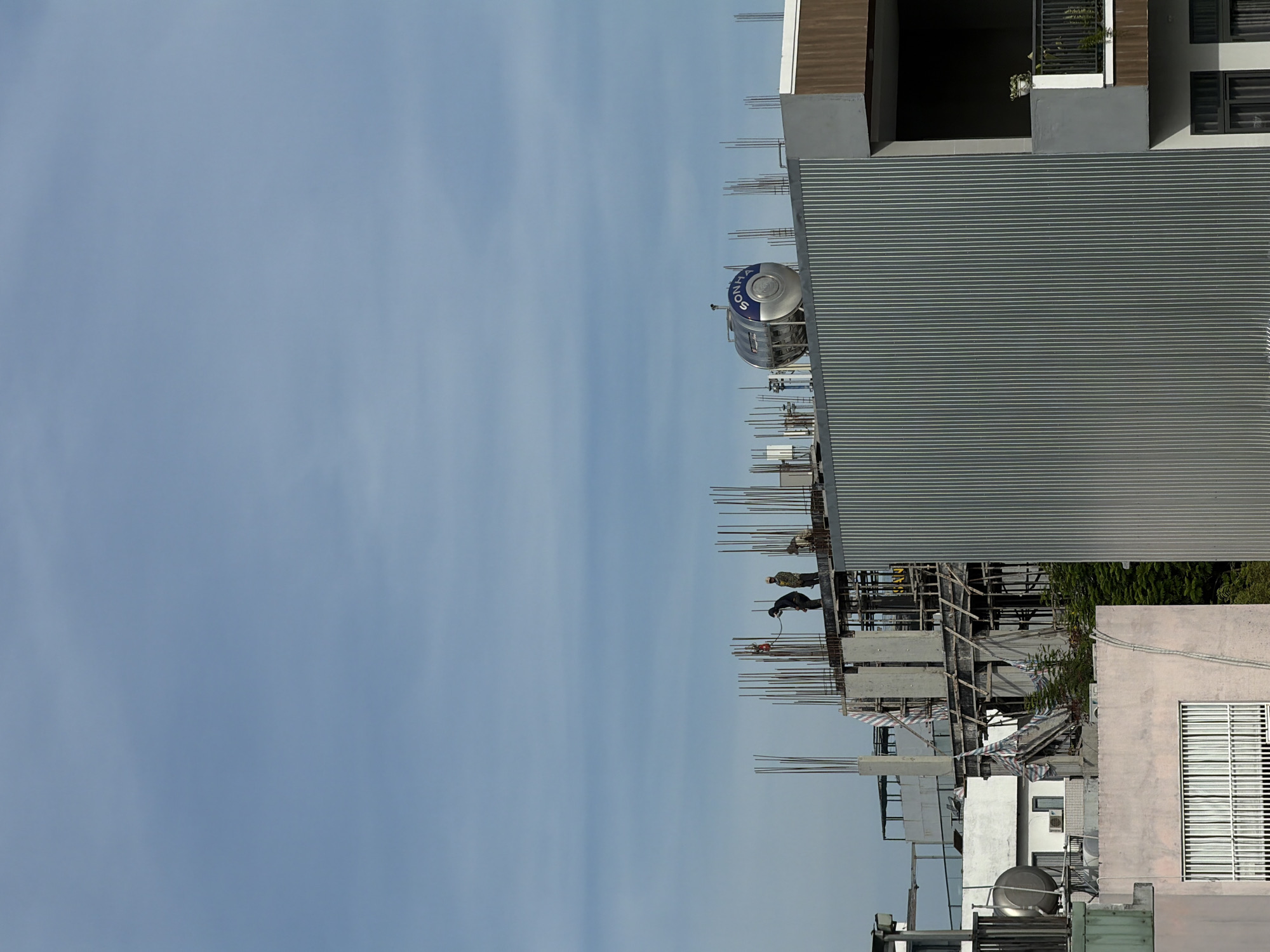 The image shows a construction site in Đà Nẵng, Vietnam. Two workers are welding metal on a high platform, with scaffolding visible. A water tank is on top of a nearby building. A partially constructed building is on the right, with metal siding and rebar extending upward. The sky is light blue with some clouds. Other buildings are visible in the background.
