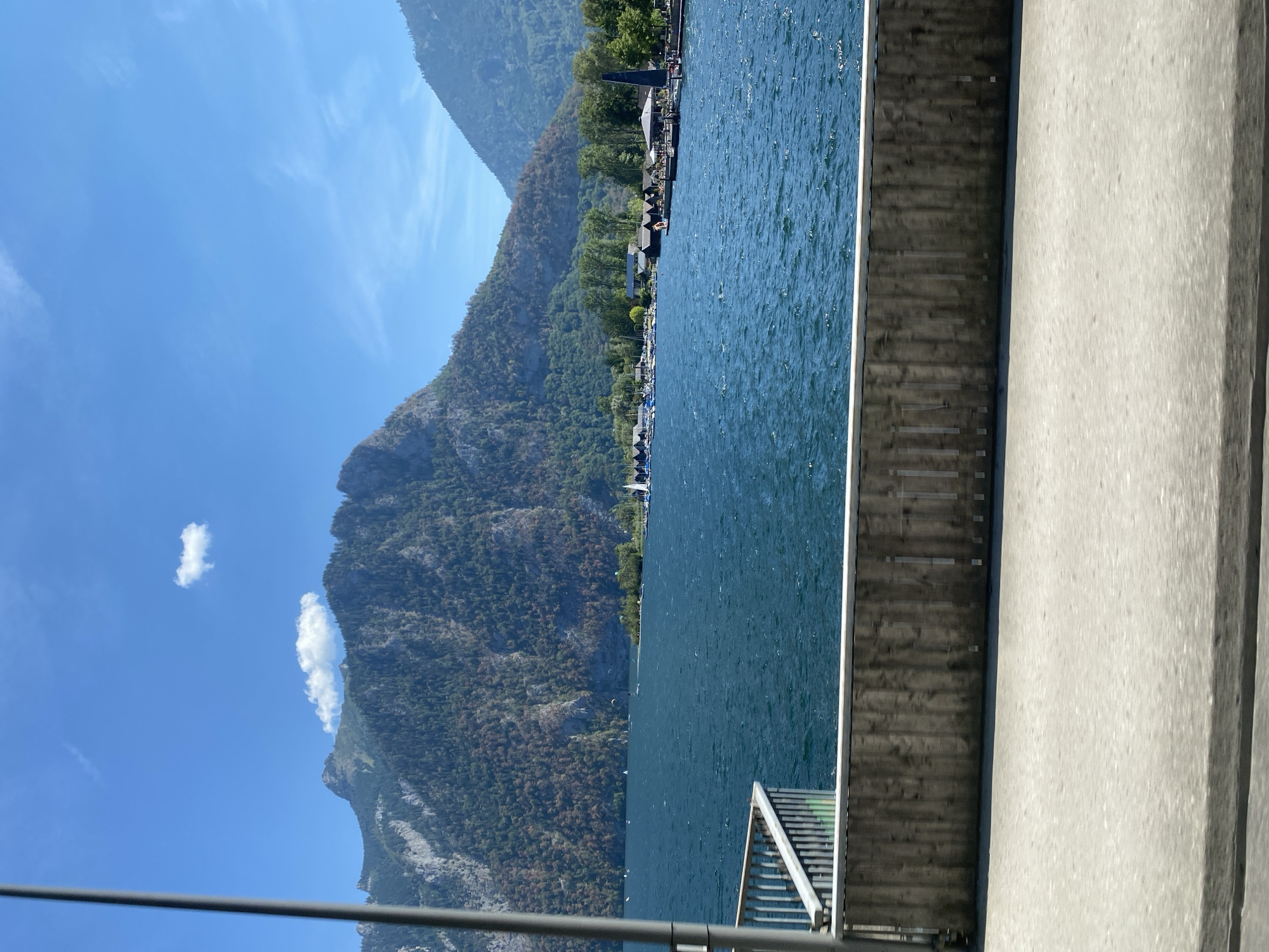 The image depicts a scenic view of Lake Traunsee in Ebensee, Austria, on a clear day with a bright blue sky and a single fluffy white cloud. The foreground features a gray concrete road or walkway, with a wooden barrier and metal railing separating it from the vibrant blue water of the lake. In the middle ground, the lake is dotted with several sailboats and a few other smaller vessels. Along the far shore of the lake, a picturesque village is nestled at the base of imposing, tree-covered mountains. The village consists of various buildings, including what appear to be boathouses or recreational structures, some with dark roofs. A prominent dark triangular structure, possibly a sail or architectural feature, stands out near the waterfront. The mountains themselves are steep and covered in dense green foliage, with some patches of exposed rock and evidence of browning vegetation on their slopes. The overall impression is one of a peaceful lakeside landscape, likely a popular tourist destination.