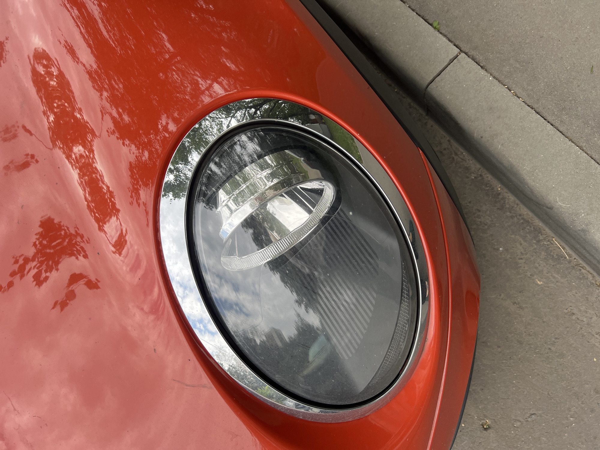 A close-up, high-angle shot captures the bright red, polished exterior of a car's headlight assembly, which is encased in a chrome trim ring. The headlight itself is dark, with a reflective surface and a ribbed pattern inside. The car's bodywork shows reflections of trees and the sky. The lower portion of the image reveals a grey concrete curb and sidewalk, with a few scattered small leaves or debris. The scene is outdoors, with daylight illuminating the car. The location of this image is not provided in the image itself.