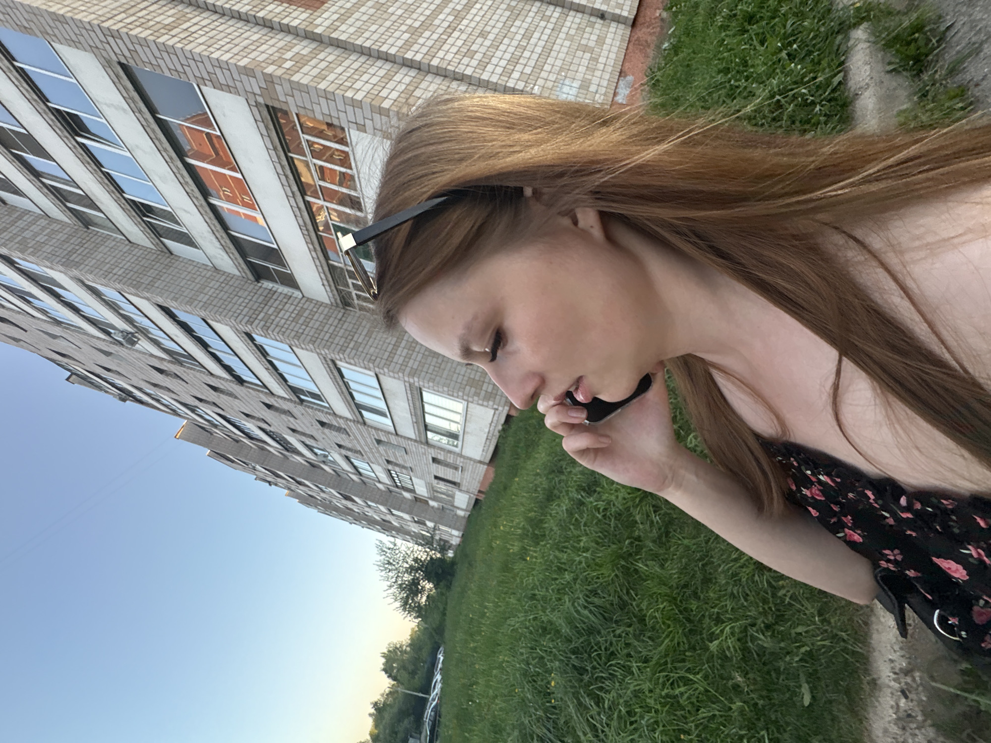 The image shows a woman speaking on a mobile phone in front of a large, residential building, likely located in Izhevsk, Russia. The building is light brick with multiple rows of windows and is angled upwards in the frame. The woman is in the foreground, facing left, holding her phone to her ear. She has long brown hair and is wearing sunglasses on her head. She's wearing a dark dress with a floral pattern. Behind her is a grassy area and possibly parked cars in the background. The sky is visible in the upper left corner.