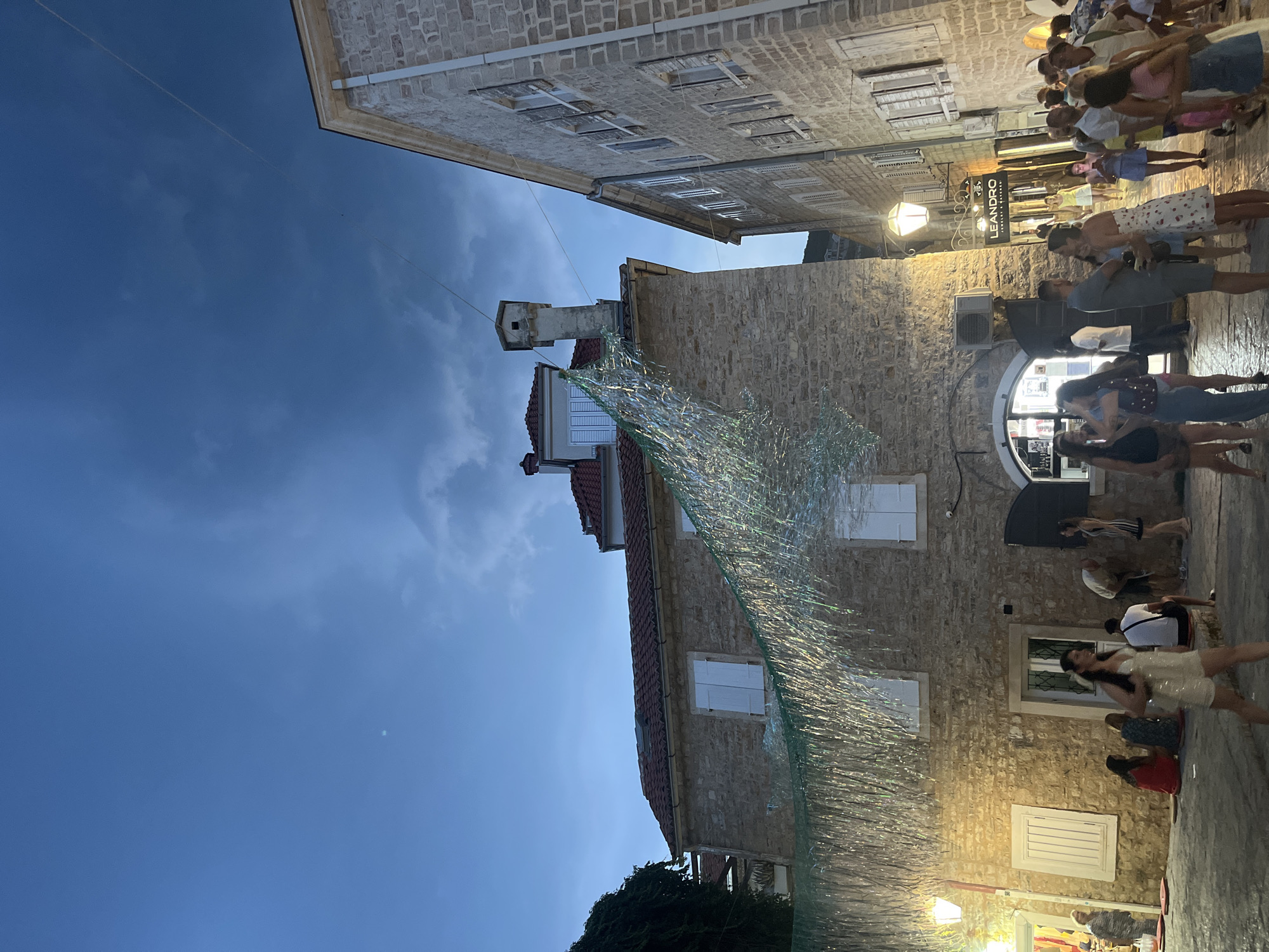 The image captures a street scene in the old town of Budva, Montenegro, during twilight. The architecture is characterized by stone buildings with red tile roofs, and the sky is a deep blue with some wispy clouds. A decorative, shimmering garland hangs across the street, strung between the buildings.
In the foreground and midground, a group of people are gathered. Some are standing, while others are seated on cushions on the cobblestone ground. They appear to be casually socializing, with some holding phones. One woman in a white sparkly outfit is walking towards the camera. The lighting suggests it is evening, with streetlights illuminating some areas and casting warm glows.
A sign for "LEANDRO JEWELRY & WATCHES" is visible on a building in the mid-right of the frame. The overall atmosphere is relaxed and social, with the historic setting of Budva's old town providing a picturesque backdrop. The image is taken from a low angle, looking upwards towards the buildings and sky.