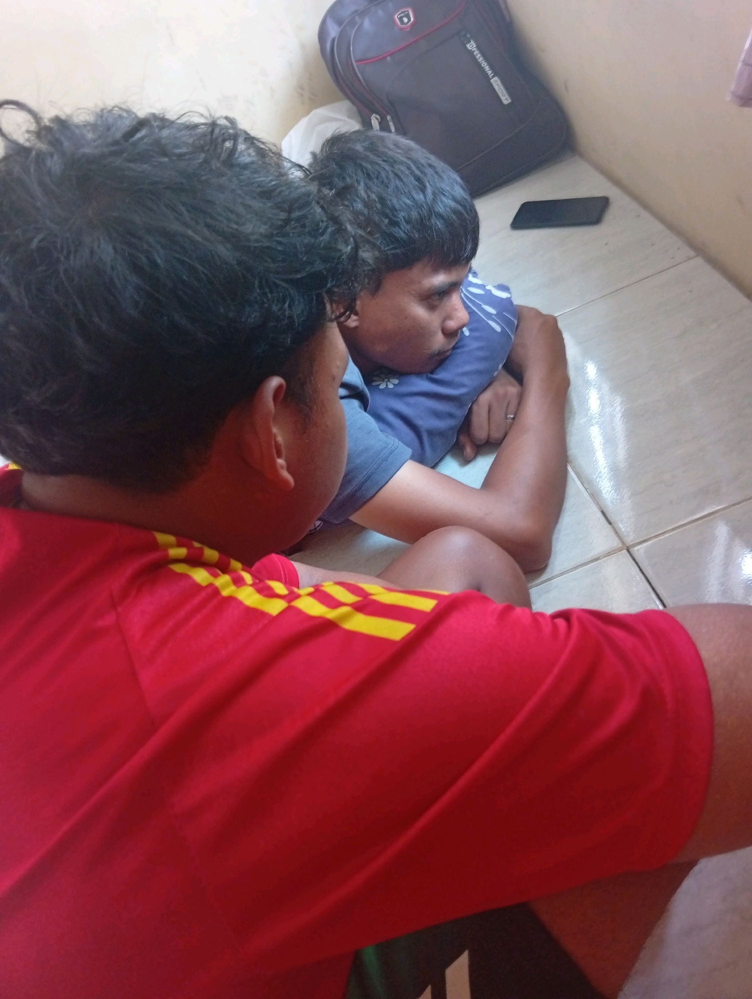 In the city of Kualu, Indonesia, two individuals are positioned on a tiled floor. The person in the foreground, facing away from the camera, wears a red t-shirt with yellow stripes on the sleeves. The second individual, facing left, is lying down with their head resting on a blue pillow, wearing a blue t-shirt and a ring on their left hand. To the right, a black backpack rests against a pale wall, and a dark smartphone is placed on the floor near the backpack. The environment appears to be an indoor space with tiled flooring and a light-colored wall.