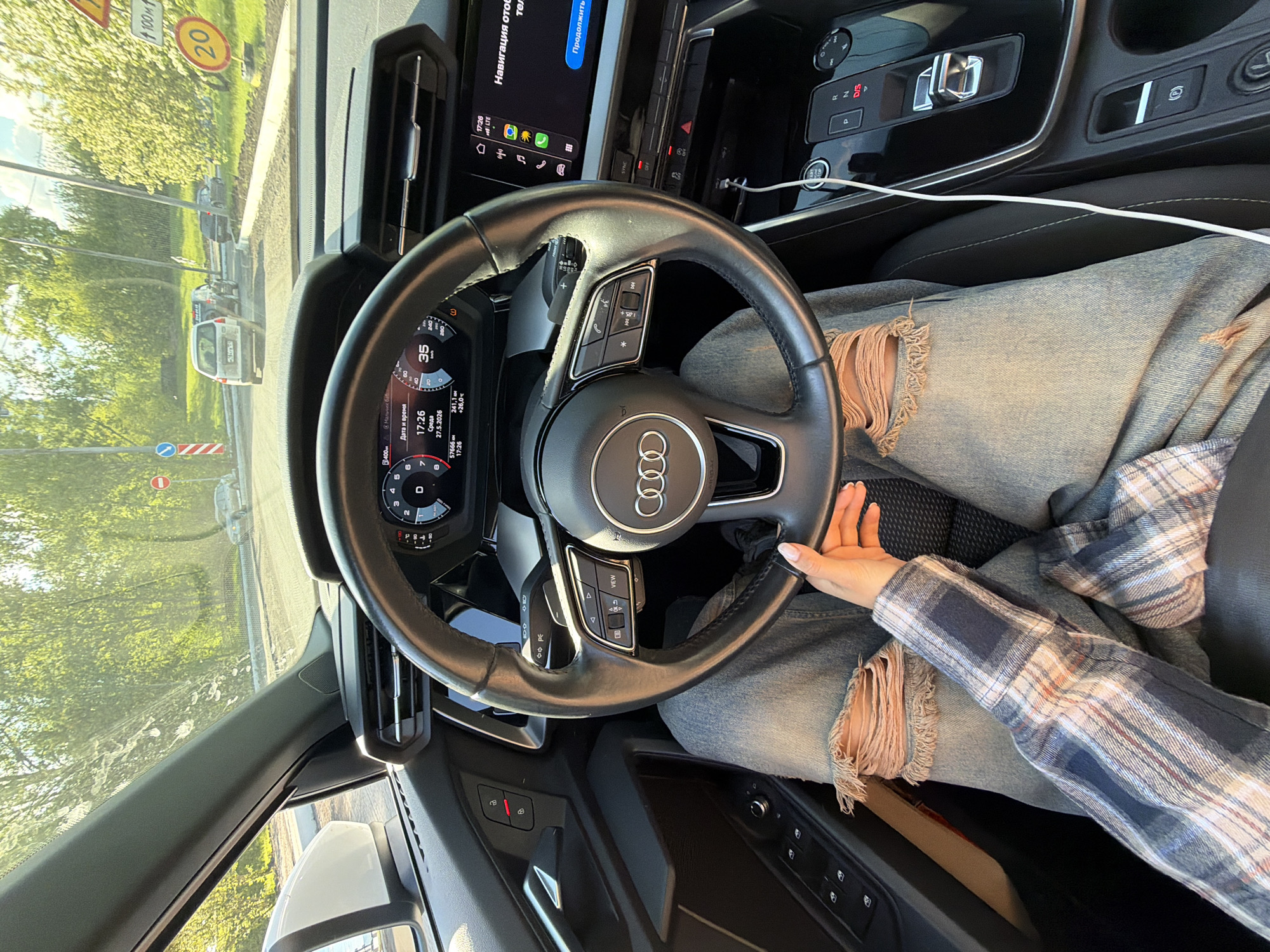 Here is a detailed description of the image:
**Scene:** The image captures the interior of a car, specifically the driver's seat area. The viewpoint is from slightly above and behind the steering wheel.
**Objects:**
* **Car Interior:** A black leather steering wheel with the Audi logo prominently displayed. Behind the steering wheel is the car's dashboard. A screen displays information, including the date and time. The center console includes gear shift buttons. A white charging cable is visible. A view of the outside, showing a road with cars and greenery. A speed limit sign of "20" and another with the number "100 м."
* **Clothing:** The driver is wearing ripped light blue jeans and a plaid shirt in light colors.
* **Person:** The hand and leg of an individual.
**Environment:** The presence of the road and trees visible through the windshield suggests an urban or suburban driving setting.
**Details from the Screen:**
* Date and Time: 17:26 on Wednesday, May 27, 2026
* Connectivity: The "LTE" icon indicates network connectivity.
* Navigation: There is a display related to navigation.
**Location:** Based on the text "Мальчик" on the dashboard screen and other elements, the location is possibly the city of Krasnoyarsk, Russia.