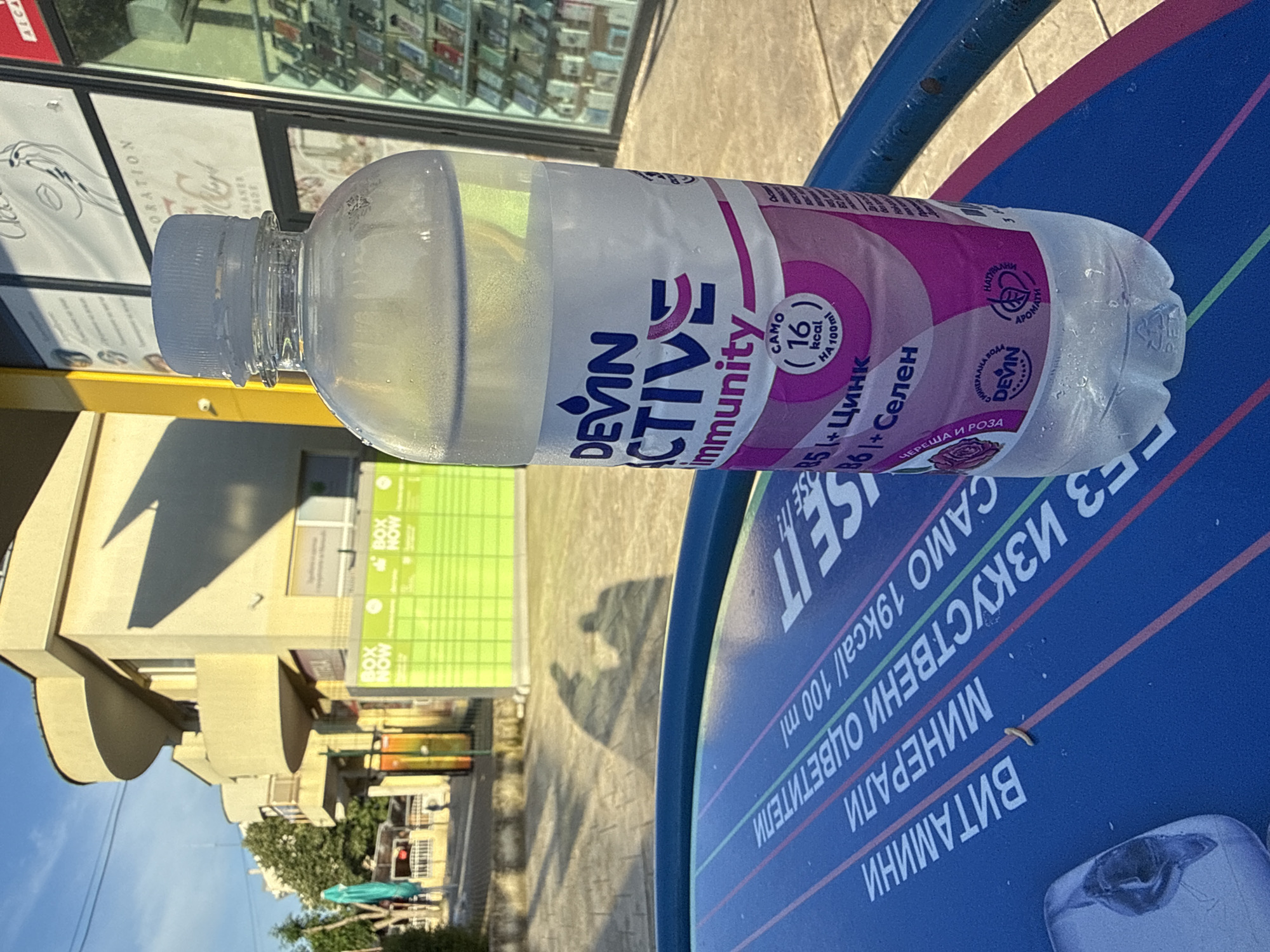 An outdoor, slightly tilted shot captures a plastic bottle of Devin Active Immunity drink lying on a blue surface. The bottle, partially filled with a clear liquid, has condensation on its exterior and a light blue cap. Its label is white and pink, prominently displaying "DEVIN ACTIVE Immunity." Further text on the label indicates "CAMO 16 kcal НА 100ml" (ONLY 16 kcal per 100ml), "B5 + Цинк" (B5 + Zinc), "B6 + Селен" (B6 + Selenium), and "ЧЕРЕША И РОЗА" (Cherry and Rose), along with "МИНЕРАЛНА ВОДА" (Mineral Water) and "НАТУРАЛНИ АРОМАТИ" (Natural Aromas).
The blue surface beneath the bottle, possibly a table or bench, features white and pink stripes and additional text in Cyrillic, including "ЕЗ ИЗКУСТВЕНИ ОЦВЕТИТЕЛИ" (NO ARTIFICIAL COLORS), "CAMO 19kcal/100ml" (ONLY 19kcal/100ml), "МИНЕРАЛИ" (MINERALS), and "ВИТАМИНИ" (VITAMINS). A small, worm-like insect is visible on this surface.
The background reveals an urban setting with several buildings under bright daylight, indicated by strong shadows, including a notable shadow resembling a person on the ground. A bright yellow structure is in the mid-ground. Green signage with "BOX NOW" repeated is visible on one of the buildings, and further text like "CORATION" and "ALCA" can be partially seen on a glass storefront. Green foliage from trees is visible further back. The scene is located in Shumen, Bulgaria.