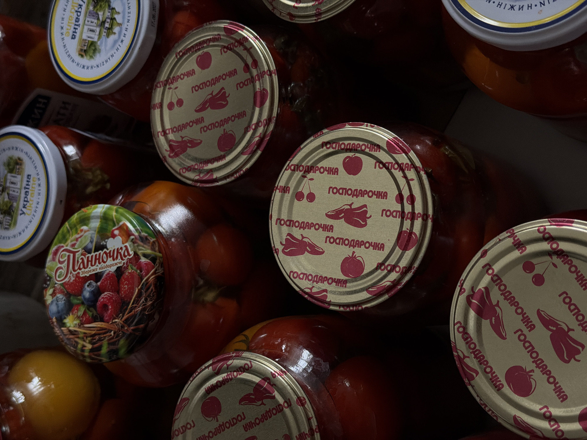 The image shows a collection of preserved food items in glass jars, likely homemade or from a small producer. Several jars are topped with metallic lids, some featuring embossed designs of fruits like cherries and peppers, and text that appears to be in a Cyrillic script, possibly indicating "Gospodarochka" and possibly "tomatoes." One jar has a colorful lid depicting berries such as raspberries and blueberries, along with text that reads "Ланночка" (Lanochka) and "Збереже надовго" (Will preserve for a long time). Another jar lid shows an image of a building and the word "Україна" (Ukraine). The contents of the jars appear to be primarily red fruits or vegetables, possibly tomatoes or peppers, submerged in liquid. The lighting is dim, and the focus is on the jars and their lids. The overall impression is one of preserved food items, possibly for storage or sale. There is no discernible indication of the specific location within Khmelnytskyi, Ukraine, or any people present in the image.