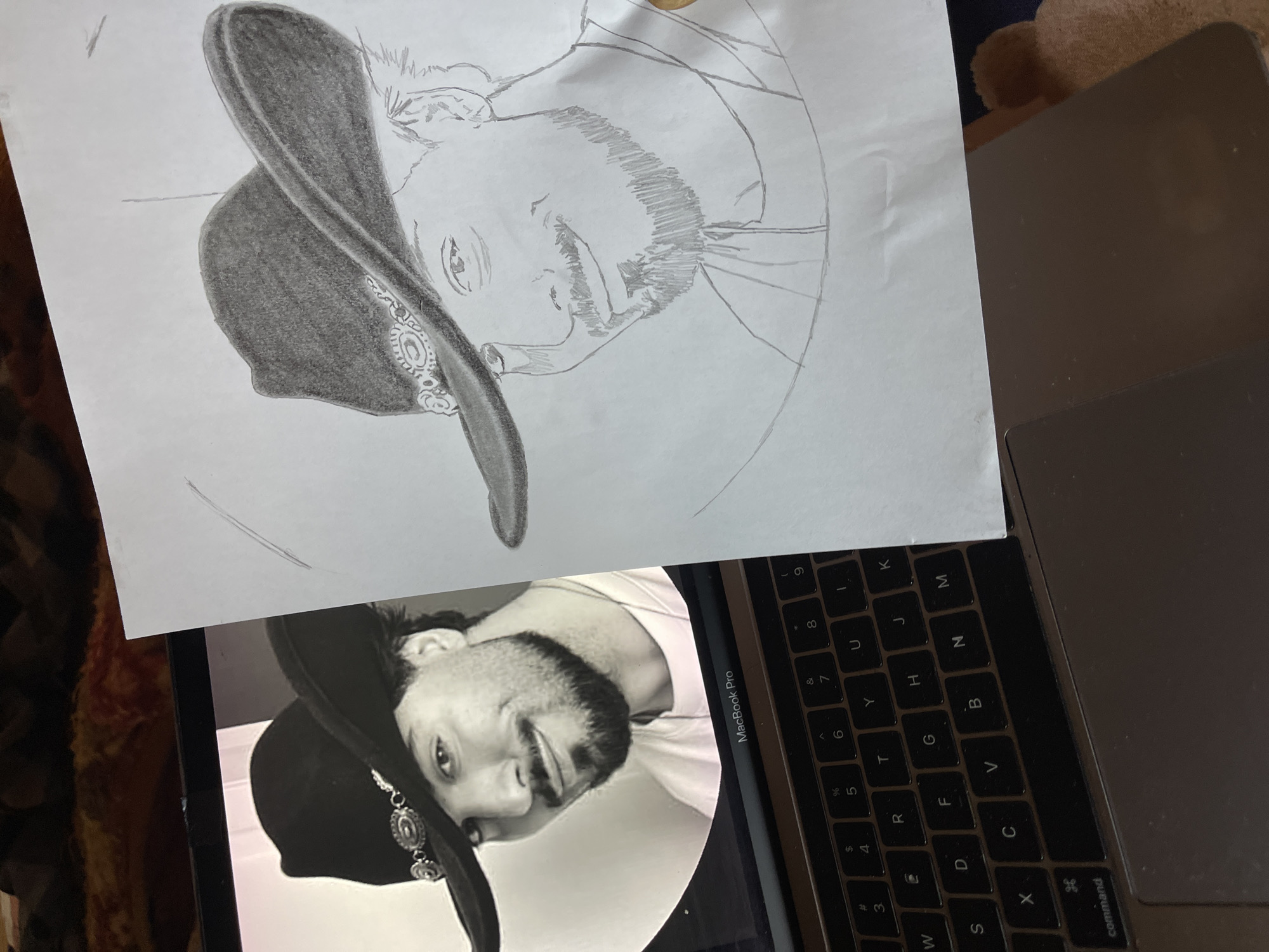 Here is a description of the image.
A photograph depicts a hand-drawn pencil sketch of a man wearing a cowboy hat. The sketch is on a piece of white paper that is resting on a laptop. The laptop screen displays the black and white photo of the same man from which the sketch was created. The laptop is a MacBook Pro with a black keyboard, and a portion of the laptop's silver casing is visible. The top of the photo contains a section of a dark brown carpet. The image is well-lit, suggesting indoor lighting. The man is depicted from the shoulders up.