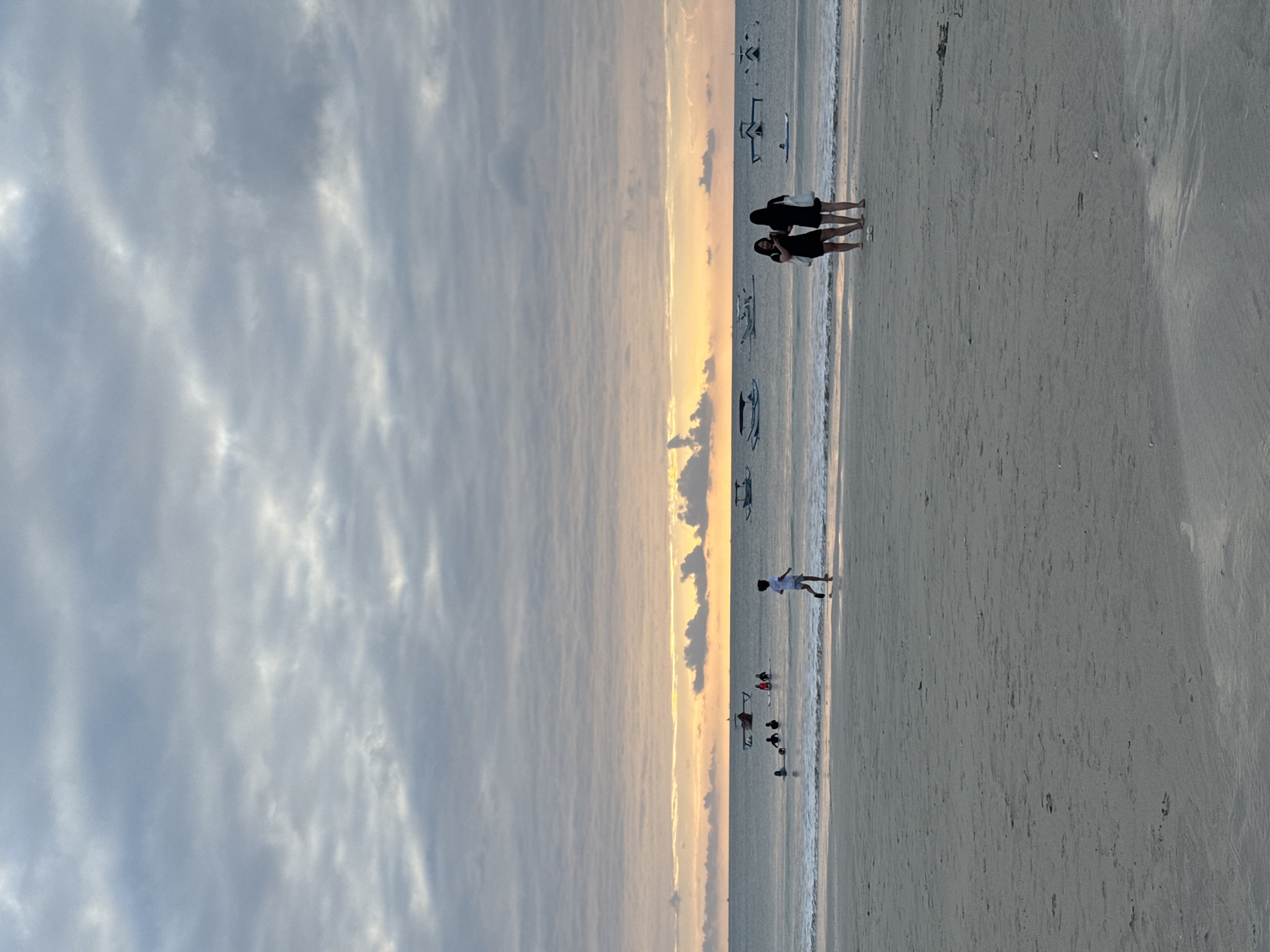This is a vertical, medium shot of a beach in Kuta Selatan, Indonesia, during a cloudy sunset. The sand is light grey and flat, with some footprints. In the distance, the ocean is calm with small waves breaking on the shore. Several boats are visible on the water, some with people in them. The sky is filled with grey clouds, with a band of orange and yellow light near the horizon, indicating the setting sun.
In the foreground, a lone figure, likely a child, is running across the sand. Further back, two women stand on the beach facing the water, each carrying a bag. In the mid-ground, a group of people are in the water, some appear to be on surfboards.