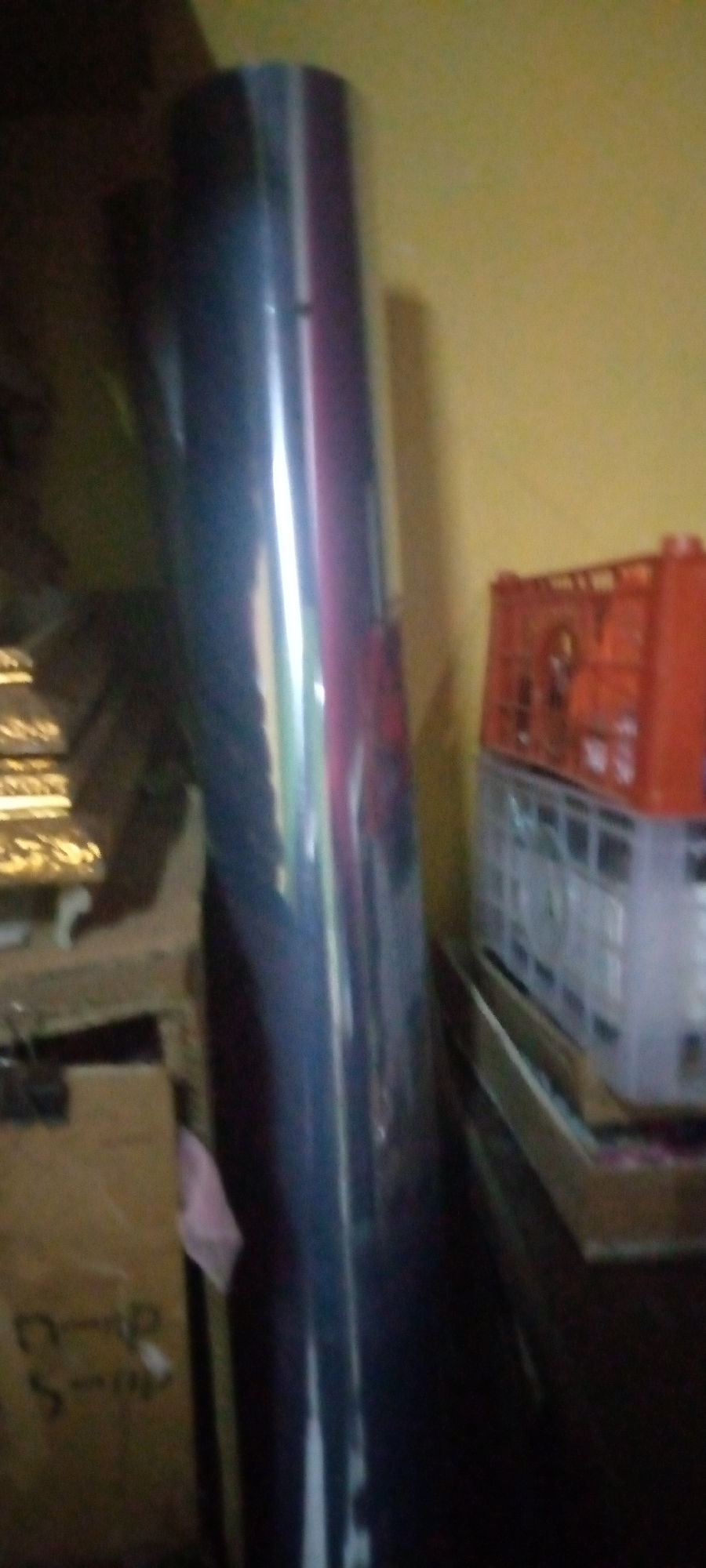 In Sumberwono, Indonesia, a blurry, dimly lit indoor scene captures several objects. Dominating the foreground is a tall, cylindrical object with a reflective, dark surface, possibly a roll of material. To its right, a bright orange crate sits atop a clear plastic container, both filled with what appear to be smaller, lighter colored items. Behind these, a yellow wall creates a stark contrast to the shadowy area on the left, which contains glimpses of what might be stacked items or boxes. The overall impression is of storage or a cluttered workspace.