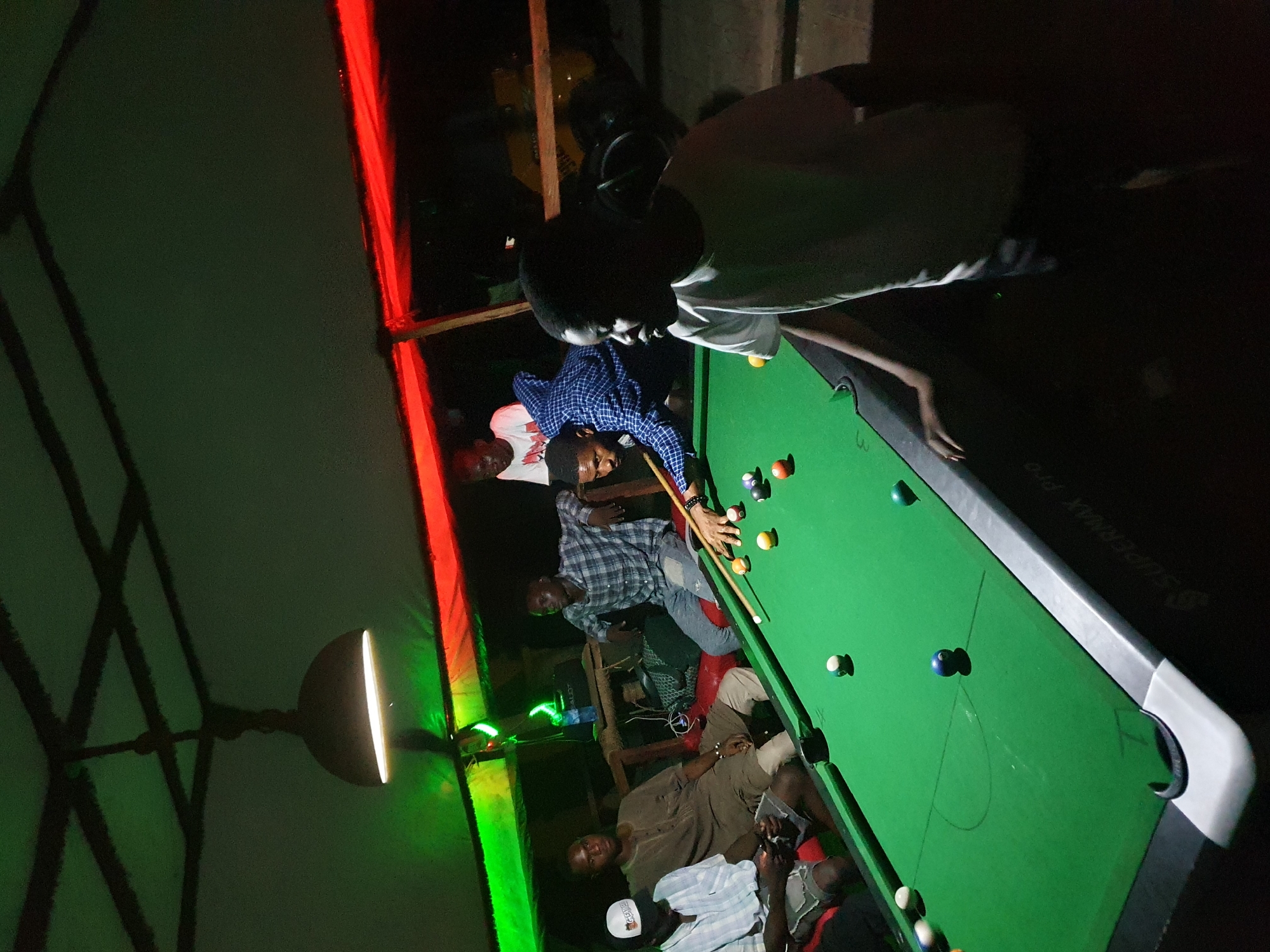 The image captures a casual, dimly lit evening scene featuring several men gathered around a pool table, likely in an indoor or covered outdoor setting. The picture is rotated 90 degrees clockwise.
**Subjects:** There are approximately seven to eight men present. One man, wearing a blue and white checkered long-sleeve shirt, is actively playing pool, leaning over the table with a cue in hand. Another man in a white t-shirt stands near him, observing. At the far end of the table, a man in a light-colored top stands, watching the game. Other men are seated or standing around the table, engaging in conversation or observing the game. One man in a brown traditional garment is seated, another in a striped shirt and a white baseball cap.
**Objects:** The central object is a green-felted pool table with multiple colored and white pool balls scattered across its surface. A pool cue is visible in the hands of the player. Artificial lighting comes from red and green LED strip lights mounted along a structure on the left side of the frame, and a single downward-facing lamp casting light onto a part of the tent-like ceiling structure. Some chairs or stools are present for the seated individuals.
**Activity:** The primary activity is a game of pool, with one man taking a shot while others observe and socialize. The atmosphere appears relaxed and recreational.
**Notable details:** The scene is illuminated by artificial light, suggesting it is nighttime or a dark interior. The red and green lights create a distinct, ambient glow. The environment seems to be a simple, functional space, possibly a community center, bar, or a recreational spot in Lokoja, Nigeria. The men are casually dressed.
**Visible text:** The text "SUPERMAX PRO" is faintly visible on the grey side rail of the pool table.