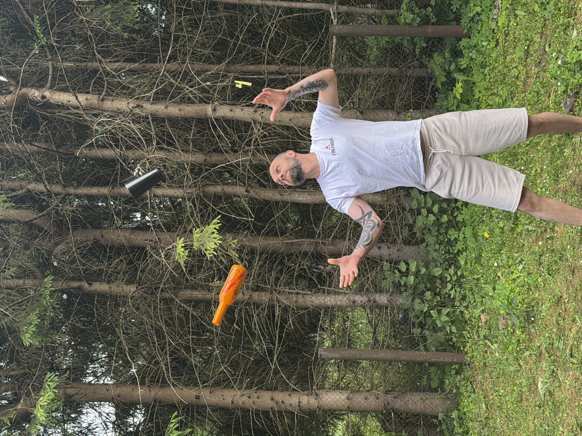 A bald, bearded man with tattoos on his arms is juggling three objects in a forest clearing. He is wearing a white t-shirt and light-colored shorts. One object, an orange bottle, is in mid-air to the left of the man. Another object, a black cylinder, is in mid-air to the right of the bottle. A third object, a yellow piece of material, is in mid-air above the man's head. The man is looking up at the objects, his arms outstretched as if to catch them. The forest clearing is surrounded by tall, thin trees with dense branches. A chain-link fence is visible in the background. The ground is covered in grass and small plants. The lighting suggests it is daytime, with the sun filtering through the trees. The location is identified as Tallinn, Estonia.