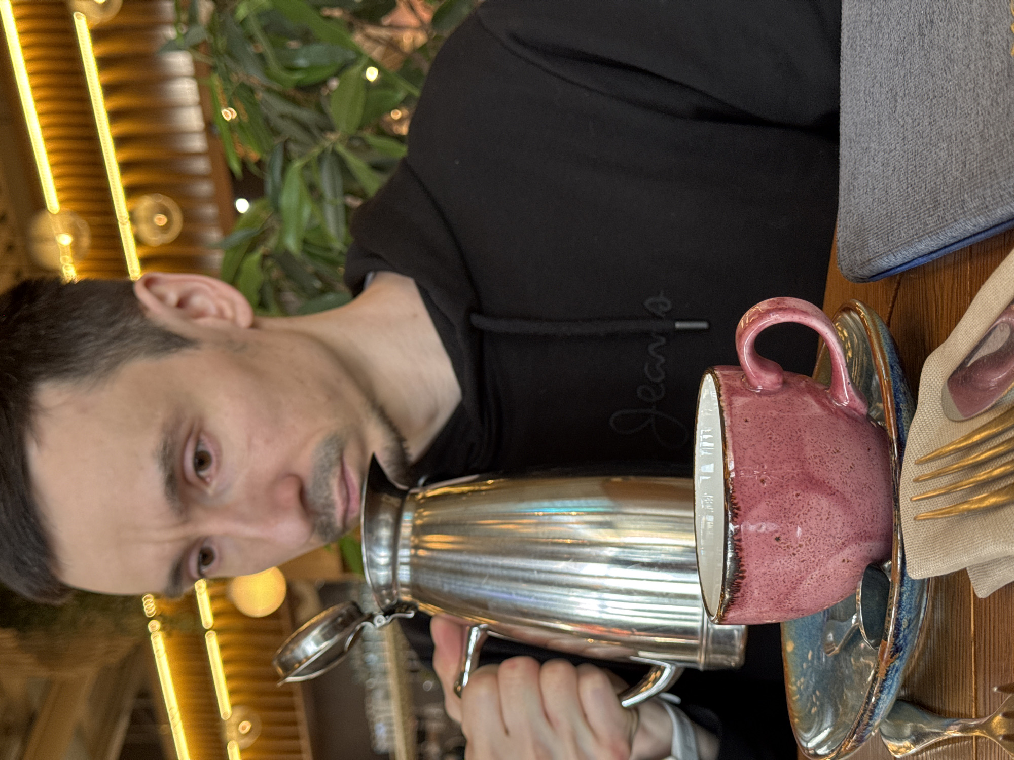 A man is holding a silver pitcher and appears to be pouring liquid into a pink and white teacup. The cup is resting on a blue and gold saucer. Next to the cup and saucer is a beige napkin with silverware. The man is wearing a black hoodie with white writing on the sleeve. The location appears to be a restaurant with decorative lights and greenery. Though the request is for a location in Sochi, Russia, there is no verifiable information in the image to suggest that it is taken there. It is therefore, not possible to include the request for the location.