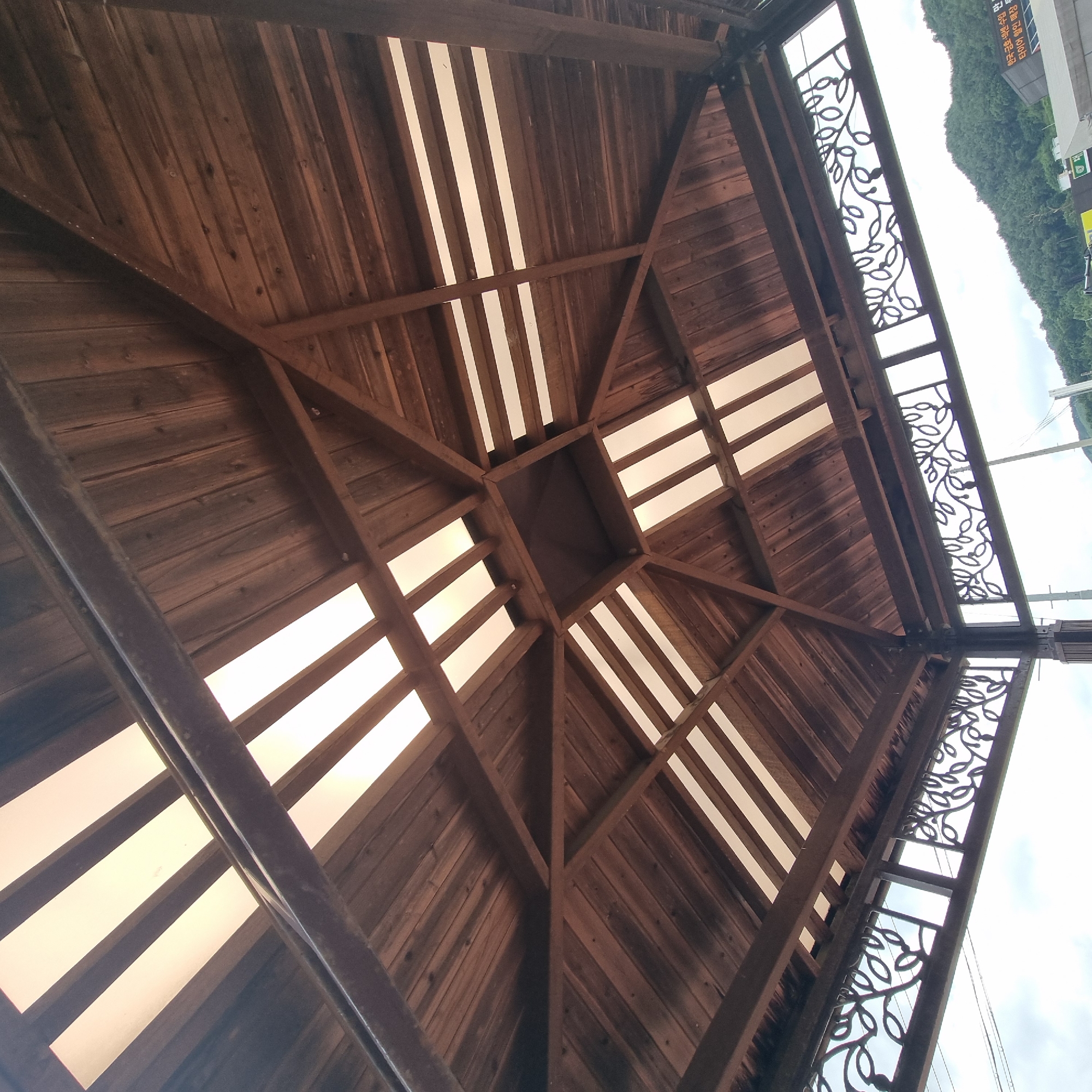 The image is a low-angle shot looking up at the wooden ceiling of an outdoor pavilion. The ceiling is constructed with dark wooden planks and features a geometric design of intersecting beams. Translucent white panels are interspersed between the wooden beams, allowing light to filter through. In the foreground, decorative metalwork with a leaf motif is visible along the edges of the pavilion. Beyond the pavilion, a backdrop of green, tree-covered hills and a cloudy sky can be seen. A sign with Korean text is partially visible in the lower right corner of the image, suggesting the location is in South Korea. While the precise location in Dunnae-myeon cannot be determined from the image alone, it depicts a common architectural feature found in public spaces or rest areas in such regions. No people or specific actions are depicted.