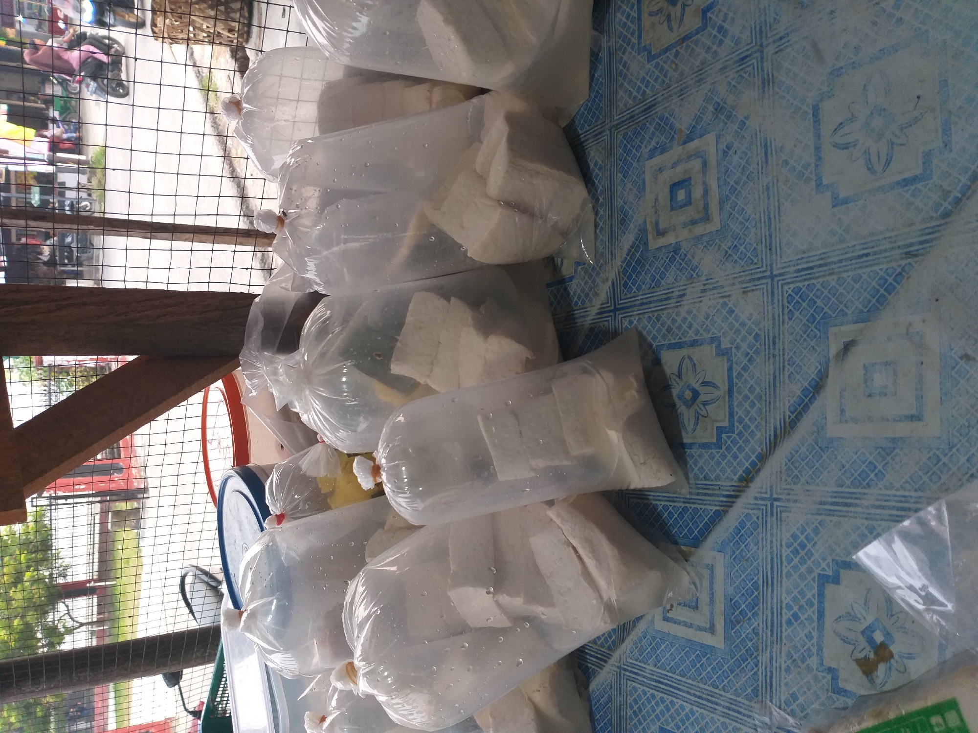The image depicts a street-level view of what appears to be a food stall or roadside market in Air Jamban, Indonesia. In the foreground, numerous clear plastic bags filled with white, cubed food items, likely tofu, are neatly arranged on a blue and white patterned floor covering. Some bags have visible water droplets on the exterior, suggesting they are packed in brine. Behind the food items, a metal fence separates the market area from a street scene. Through the fence, several motorbikes are visible, with one rider wearing a pink headscarf and another wearing a helmet. Trees and buildings line the street, indicating an urban or semi-urban environment. A wooden beam structure is also present, possibly supporting a market stall or shelter. The overall impression is one of a bustling local marketplace in Indonesia.