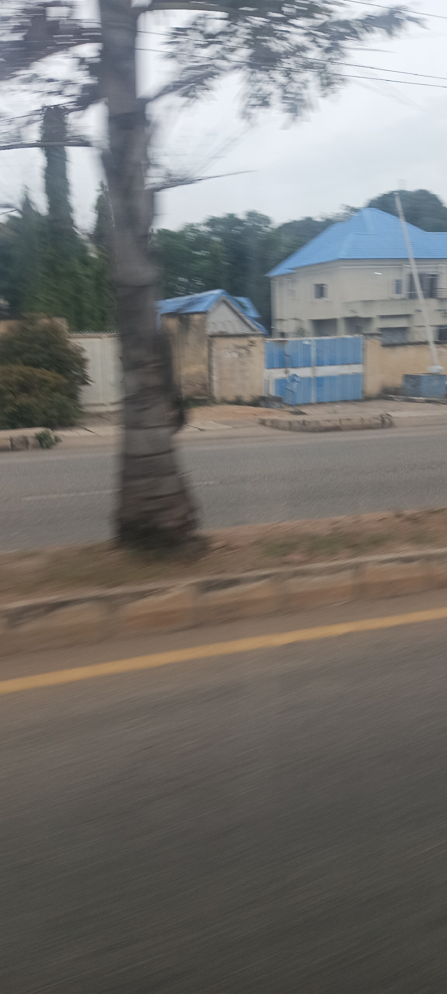 This image presents a street view, heavily affected by motion blur, suggesting it was captured from a moving vehicle in Jos, Nigeria. The blur is most pronounced in the foreground and midground, while elements in the distant background are slightly less indistinct.
In the immediate foreground, a substantial tree trunk occupies the left side of the frame, appearing as a blurred vertical mass. Below and to its right, a paved road stretches horizontally across the lower half, marked by a faded yellow line separating lanes. Beyond the road, a low curb separates it from a strip of dry, uneven ground.
Further into the scene, a light-colored compound wall is visible, behind which a small, single-story building with a striking bright blue gabled roof stands. Adjacent to this, a pair of large, blue metal gates, featuring a horizontal white stripe, provide access to the compound. To the right, a larger, multi-story residential building with a similarly vibrant blue hip roof dominates the background. This architectural choice of blue roofs is a prominent visual detail.
The upper portion of the image is filled with a pale, overcast sky and the blurred shapes of numerous trees and foliage behind the buildings. The overall lighting suggests a cloudy daytime, without direct sunlight. There are no people visible, and no discernible text on any of the structures or signs. The heavy motion blur is the primary indicator of the activity, pointing to photography from a vehicle in transit.