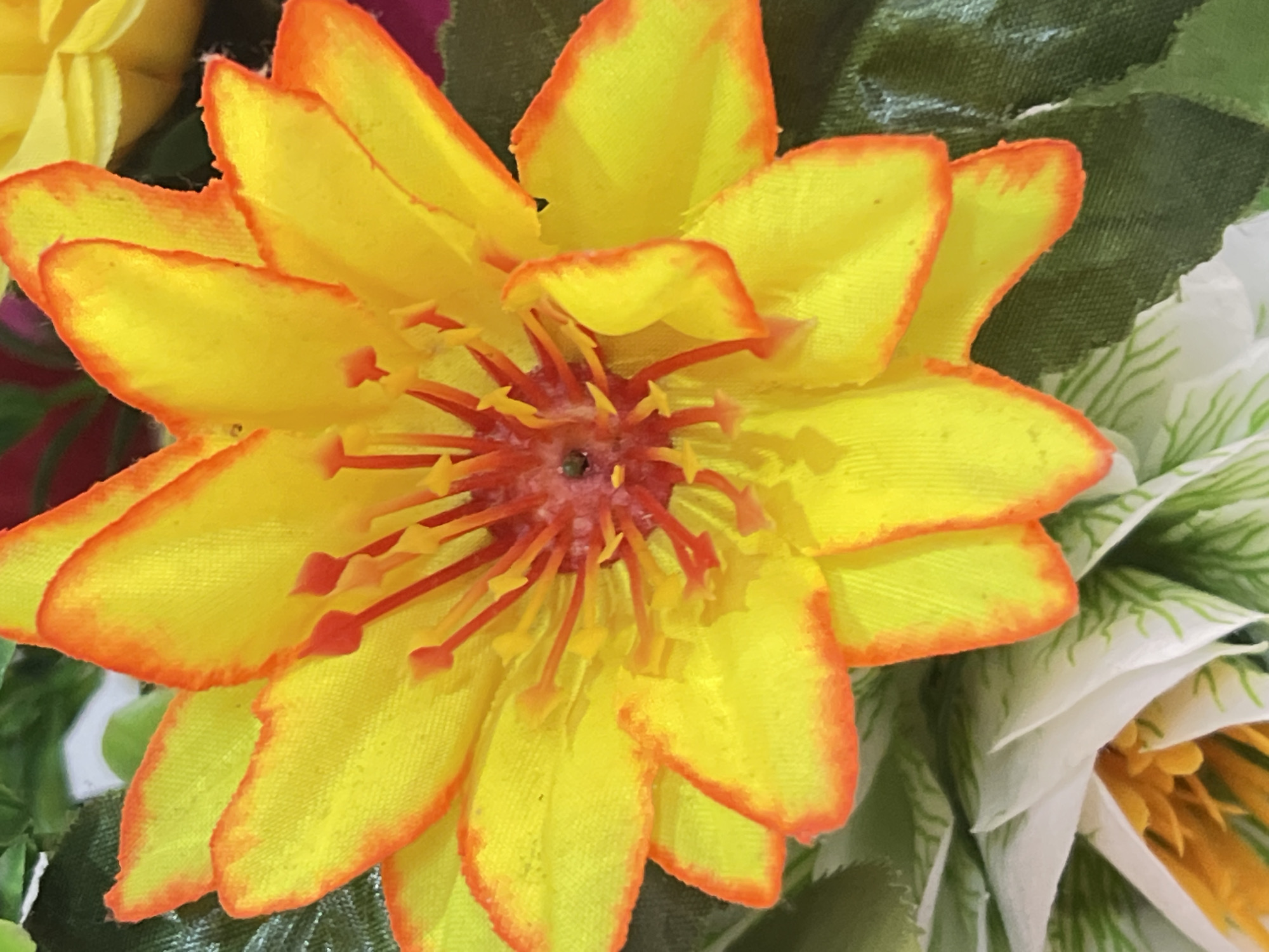 A close-up, slightly overhead shot showcases a vibrant yellow artificial flower with a red-orange trim on its petals. The flower has multiple layers of petals, with the outer ones layered over inner ones. In the center, a cluster of red stamen-like structures with yellow tips are visible. The flower appears to be part of a larger arrangement, with hints of green leaves and other artificial flowers, some white with green veins and others yellow, visible in the blurred background. The lighting is bright, highlighting the textured fabric of the petals. This image was taken in Denpasar, Indonesia.
