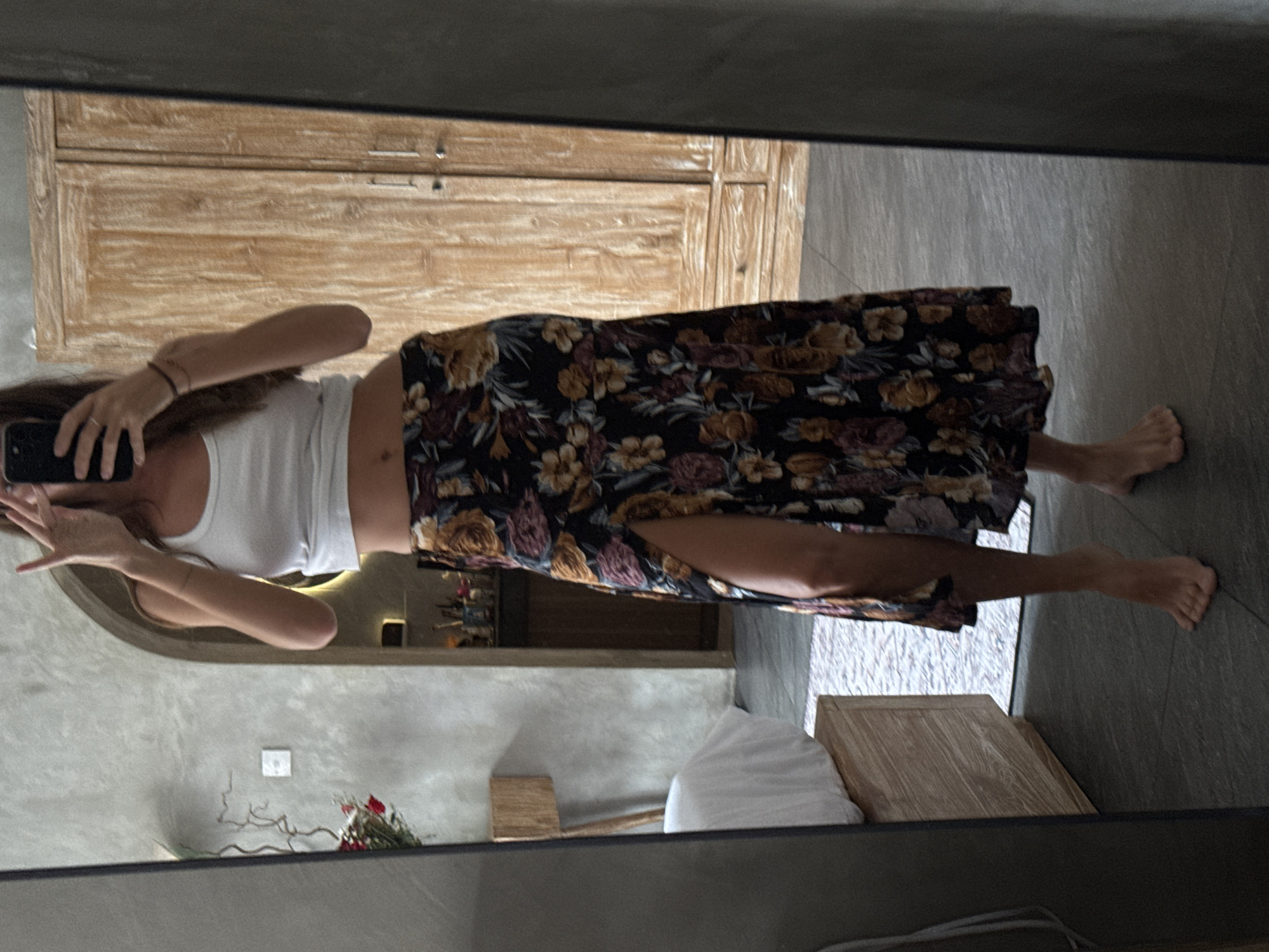 A woman is taking a mirror selfie in what appears to be a bathroom setting, reflected in a large mirror. The woman is wearing a white tank top and a floral print skirt with a slit on the side. She is holding a phone up to take the picture. The bathroom features a wooden cabinet or dresser, a white pillow on a wooden structure, and a grey wall with a white electrical outlet. There is also a small floral arrangement near the wall. Based on the interior design and the style of the furniture, it is possible that this scene is located in North Kuta, Indonesia, as the overall aesthetic aligns with the region's design preferences.