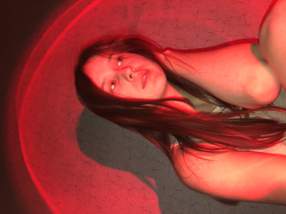 A single young female is depicted indoors, illuminated by strong red light emanating from an unseen source, which projects an arc of red light onto the textured wall in the upper background. The female has long, dark hair and fair complexion, wearing a light-colored upper garment. Her head is slightly tilted, and her gaze is directed towards the upper right portion of the frame. Her right arm is bent, with her hand positioned near her neck/shoulder, while her left arm is also bent, with the elbow visible in the lower right. A prominent, dark shadow resembling a human profile is cast on the textured wall behind her, positioned to the upper left of her head. The image provides no discernible features to confirm the location as the city of Фоки, Russia.