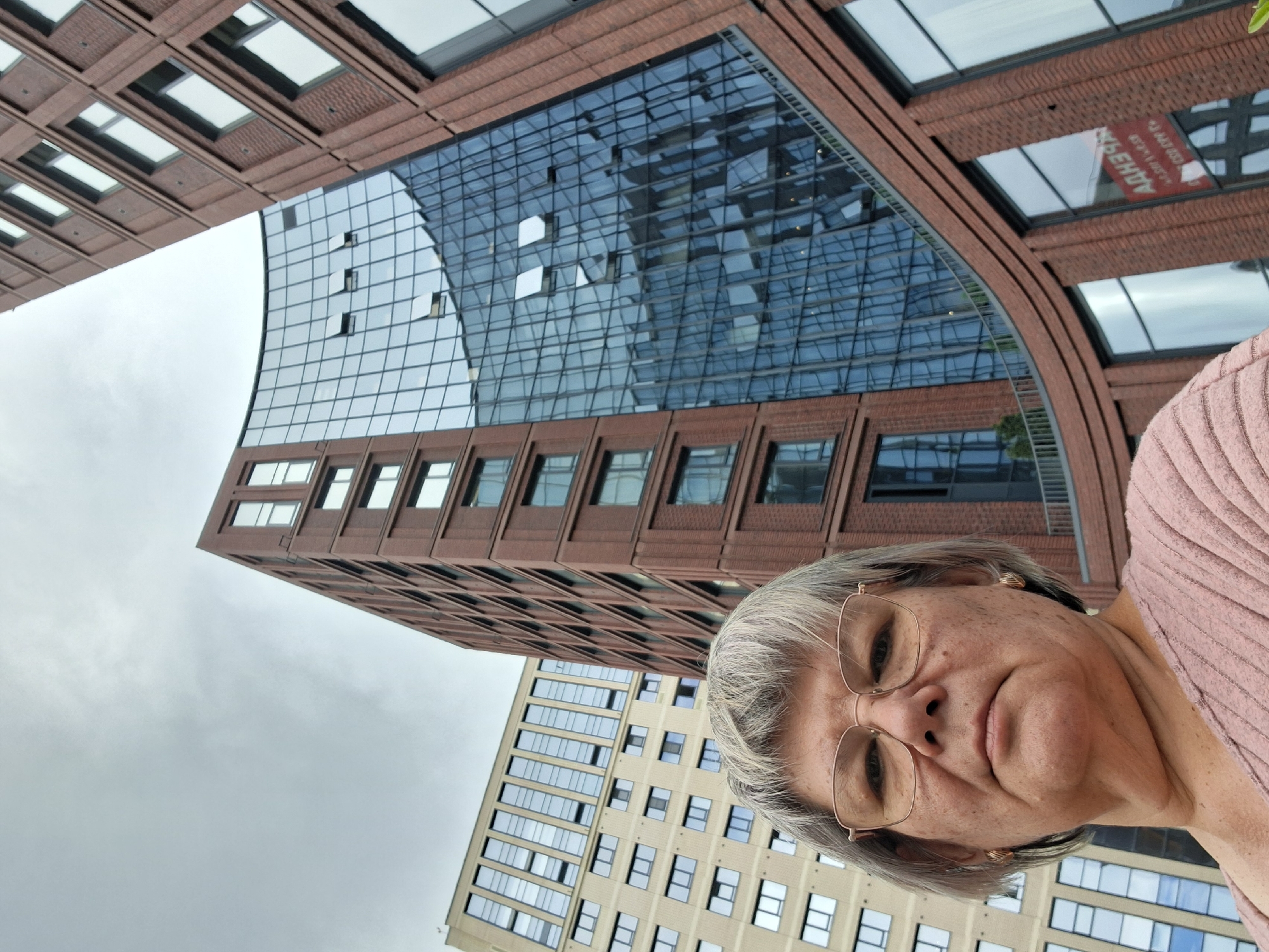 A low-angle, medium shot captures a woman from the chest up, with her face filling the lower half of the frame. She has short, gray hair, wears glasses with thin, gold-colored frames, and has on a pale pink, ribbed shirt. Her expression is neutral, and she is looking directly at the camera.
Behind her, two modern buildings dominate the background. The building on the right is a tall, brick structure with many windows, some of which are open. It curves away from the camera as it rises. To the left of this building is another, lighter-colored building with a facade composed of rows of windows. The sky above is overcast and gray.
The scene appears to be in an urban environment, likely a business district, with contemporary architecture. The red sign on the glass facade of the right-hand building has Cyrillic lettering, suggesting the location is Russia. The building on the right is the "Aurora" business center, located at 16 Komsomolsky Prospekt in Novosibirsk, Russia.