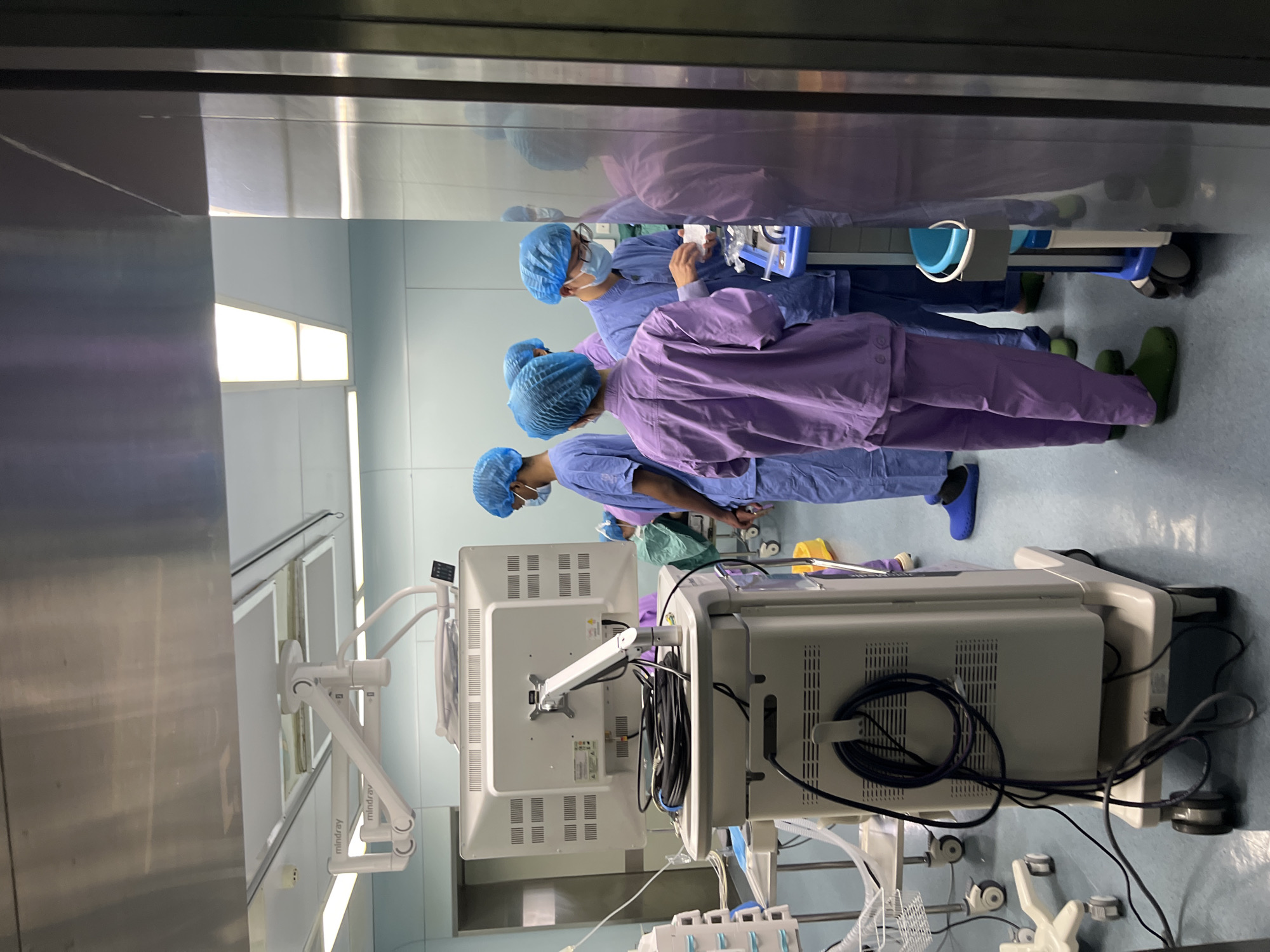 The image shows a surgical operating room. Three individuals wearing surgical scrubs, surgical caps, and face masks are present. One individual is wearing purple scrubs, while the other two are wearing blue scrubs. They appear to be involved in a surgical procedure or preparing for one. Surgical equipment including a surgical monitor, medical apparatus on wheels, and an operating light are visible. The walls of the room are light blue. The location is a hospital somewhere in the city of Yuzhong District, China.