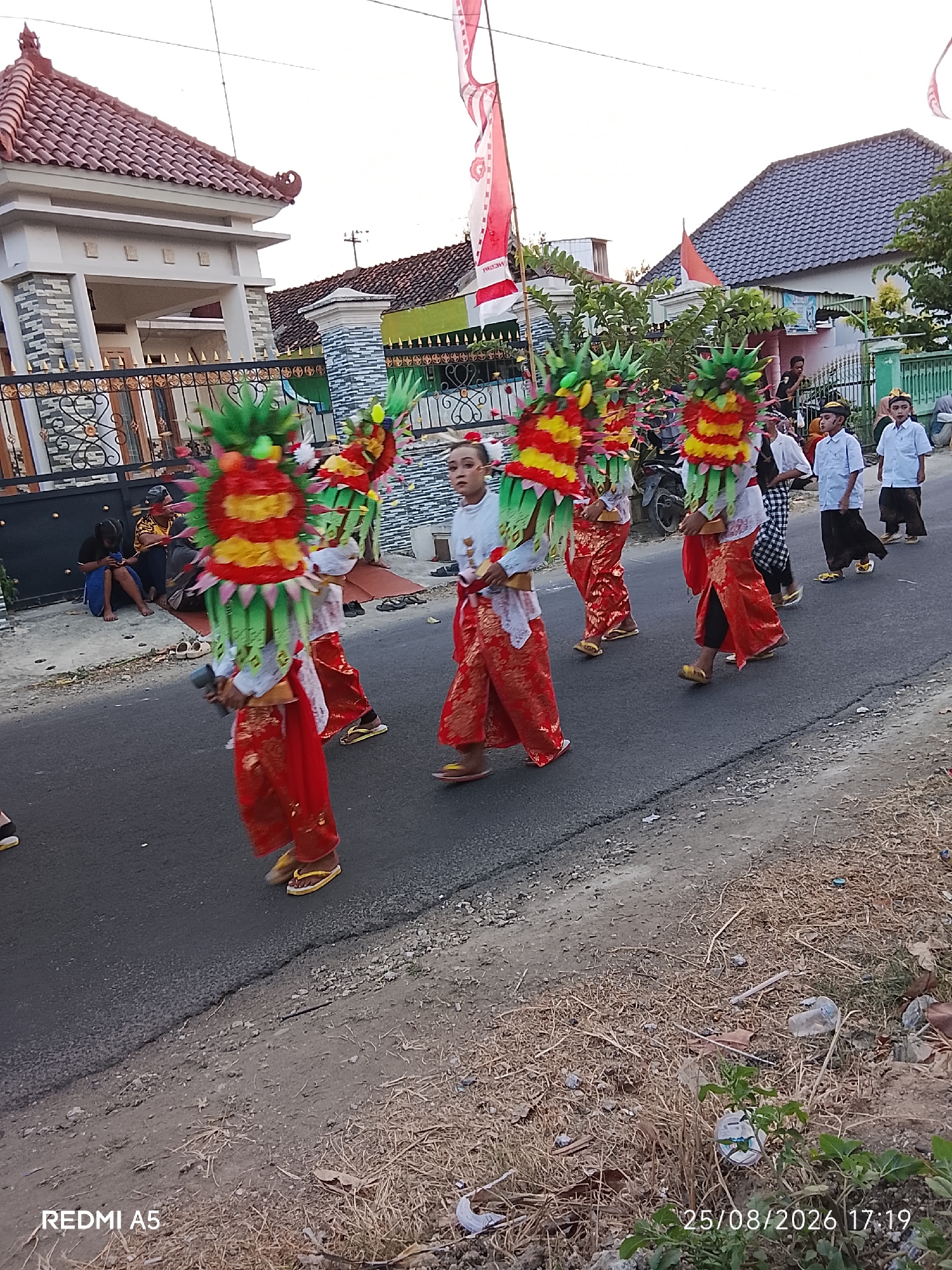 In Semanding, Indonesia, a group of individuals in traditional attire are participating in a street parade. The procession features several participants adorned in colorful, elaborate headdresses that resemble mythical creatures, with vibrant green, yellow, and red elements. They are wearing loose-fitting red trousers or skirts over white shirts, and some have yellow sandals.
Behind them, a few younger boys in white shirts and dark pants walk in a line. Spectators are visible on the side of the road, seated and standing near houses and fences. The environment consists of a paved street with a dry, grassy shoulder. Houses with tiled roofs are visible in the background, along with trees and flags, one of which appears to be the Indonesian flag. The lighting suggests it is daytime, possibly late afternoon. The overall scene depicts a cultural event or celebration taking place in a residential area.