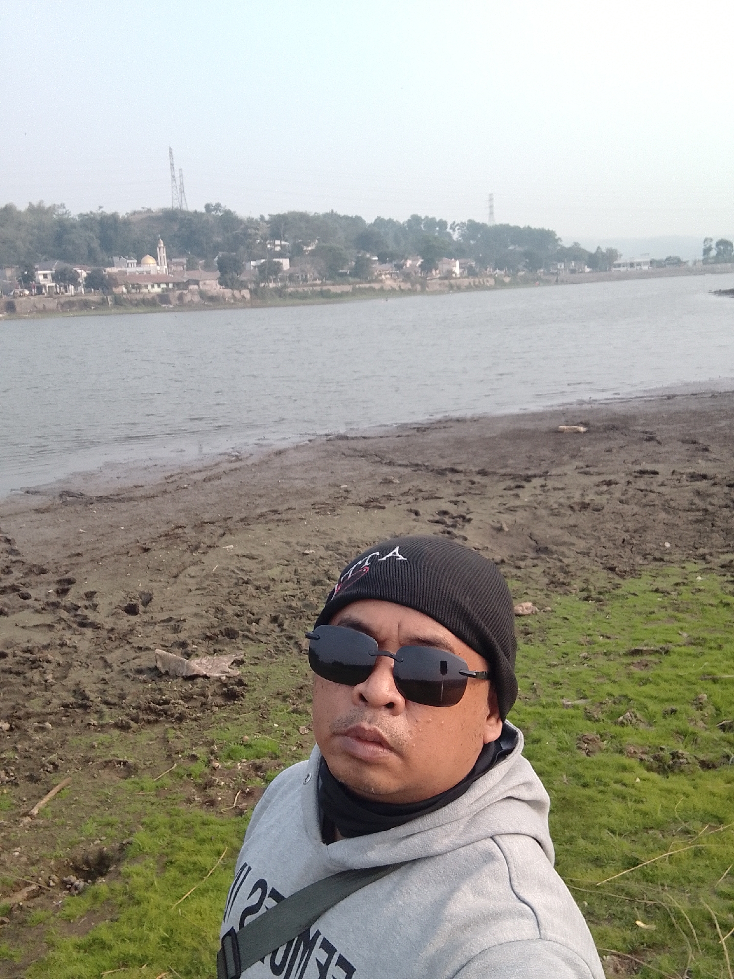 A man wearing a black beanie and sunglasses takes a selfie by a body of water. He is wearing a grey hooded sweatshirt with a dark strap across his chest. The background shows a shoreline with patches of grass and mud, leading to a wide, calm body of water. Across the water, a town is visible with buildings, trees, and a prominent golden-domed structure, possibly a mosque or church. Further inland, a large transmission tower stands on a hill. The sky is overcast, suggesting it might be morning or late afternoon. The overall scene appears to be a tranquil, rural landscape in Ciburuy, Indonesia. Visible text on the man's sweatshirt is partially obscured but appears to be in large, bold lettering.