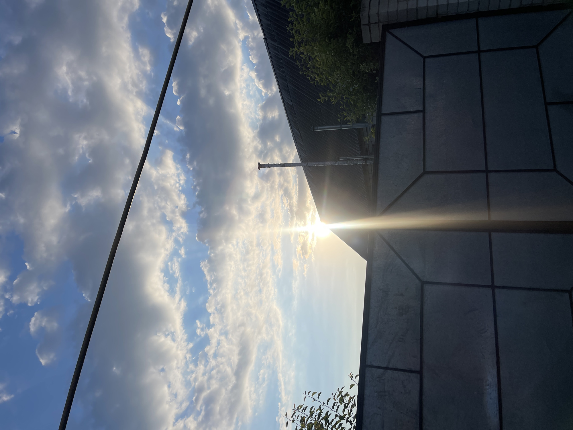 The image displays a sky filled with white and gray clouds, with patches of blue sky visible. A bright sun is visible in the sky, casting a strong glare across the image. In the foreground, a dark, angular surface is visible, possibly a rooftop or patio made of dark tiles. To the left, a dark, thin pole or wire runs diagonally across the frame. Further back, a dark, sloped roof with metal supports is visible, along with some green foliage. A brick wall is also partially visible in the upper right corner. The scene suggests an outdoor setting, likely during daylight hours due to the sun's presence. The lighting indicates it may be late afternoon or early morning, as the sun is low in the sky. There is no visible text in the image. The overall mood is somewhat dramatic due to the strong sunlight and shadows.