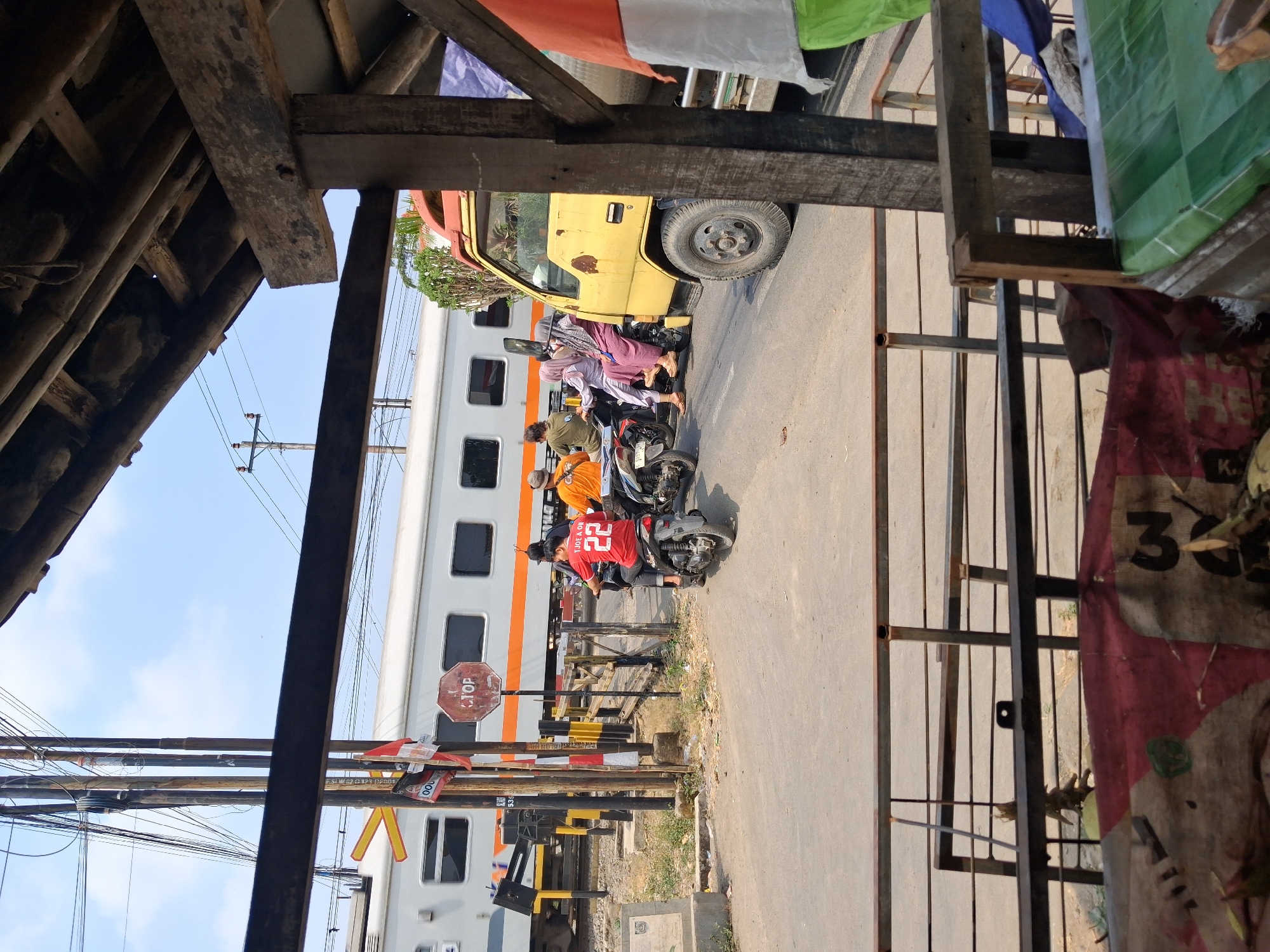 The image shows a railway crossing with a train passing by in Balapulang Wetan, Indonesia. A white train with an orange stripe is moving from left to right. Several motorcycles and a yellow truck are stopped at the crossing. On one of the motorcycles, a person wearing a red shirt with the number 22 is visible. A yellow truck is also stopped, partially obscuring the view of other vehicles. The scene is characterized by a clear blue sky and overhead power lines. In the foreground, a rustic wooden structure with a roof made of corrugated metal is visible, suggesting it might be a shelter or stall. The ground is a paved road, and there is some debris and vegetation around the crossing.