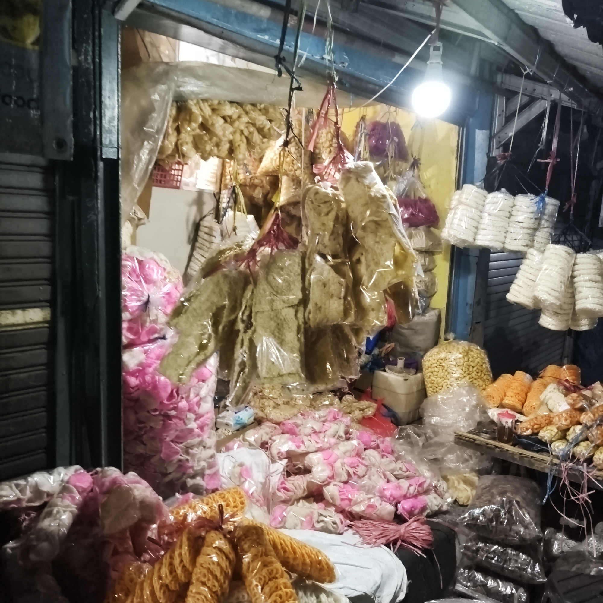 This image depicts a stall at night in South Jakarta, Indonesia, selling a variety of snacks, primarily crackers and dried goods. The stall is covered by a corrugated metal roof and illuminated by a single bright bulb. The goods are displayed in plastic bags and some are hung from the ceiling. In the foreground, there are piles of pink and white crackers, along with orange, ring-shaped crackers. To the right, stacked white crackers are visible, along with a large bag of small, yellow, bean-like snacks. Behind these, larger, flattened, dried snacks are hanging in plastic wrap. The scene appears to be an outdoor market stall, likely a common sight in the bustling urban environment of South Jakarta. There are no people visible in the image.