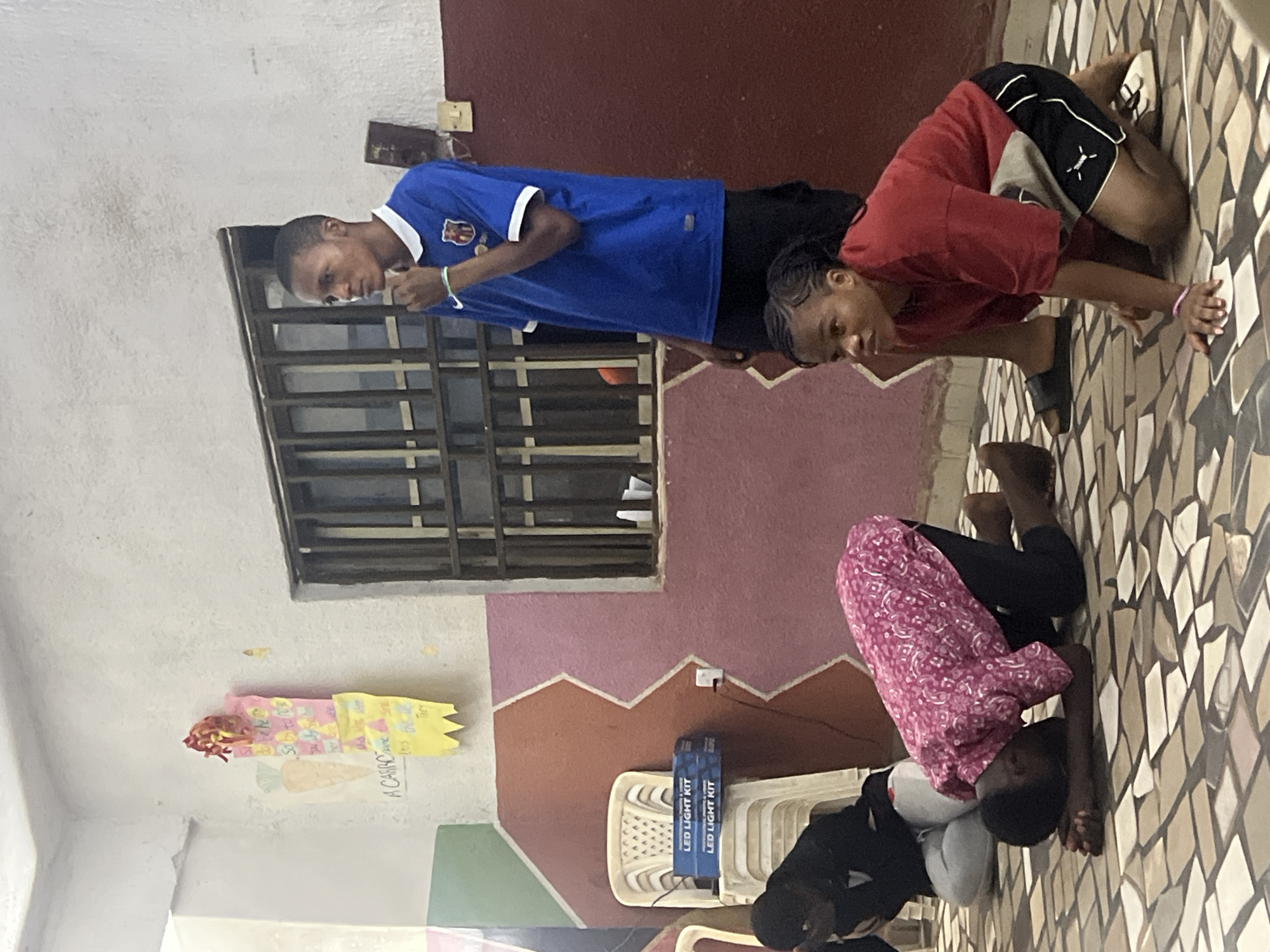 In Nkpor, Nigeria, a group of young people are engaged in what appears to be a religious or spiritual activity. The scene is indoors, with a tiled floor and walls painted in a combination of pink and reddish-brown colors, featuring a zig-zag pattern. A barred window is visible on the right side of the frame.
In the foreground, two young women are kneeling on the floor. The woman on the left is wearing a black top and white pants, with her head bowed. The woman in the center is wearing a pink patterned shirt and black pants, also with her head down and hands resting on the floor. To the right, a young woman in a red t-shirt and black shorts is also kneeling, looking upwards and to her left.
Standing behind the kneeling individuals is a young man wearing a blue and white soccer jersey and black shorts. He is looking to his right, with his hand near his chin.
Various objects are present in the scene. On a stack of white plastic chairs to the left, two boxes labeled "LED LIGHT KIT" are visible. A piece of artwork, possibly a child's drawing featuring a carrot and text, is taped to the wall on the upper left. Electrical outlets and a light switch are also visible on the walls. The overall impression is that of a gathering in a community space or a place of worship.