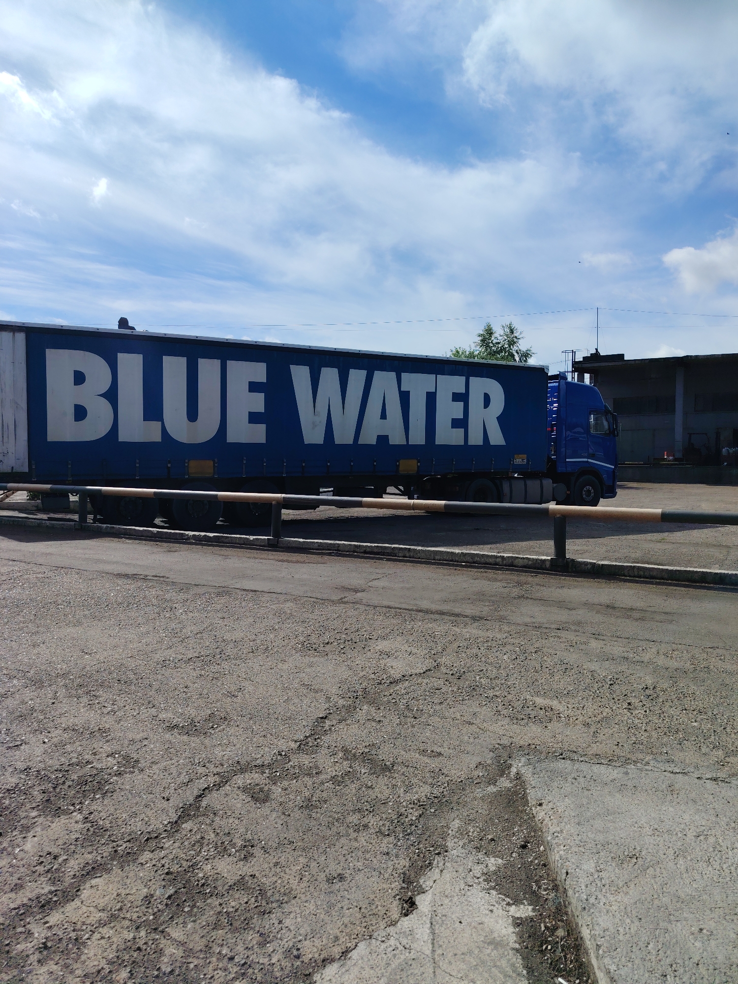 A blue semitrailer truck with the white text "BLUE WATER" on its side is parked on a paved lot. The truck's trailer is white with a blue tarp and the text is prominently displayed in large white letters. The truck cab is blue. In the background, there is a building with a dark roof and a cloudy blue sky overhead. There are several black and orange striped barriers in front of the truck. The ground in the foreground is rough asphalt.