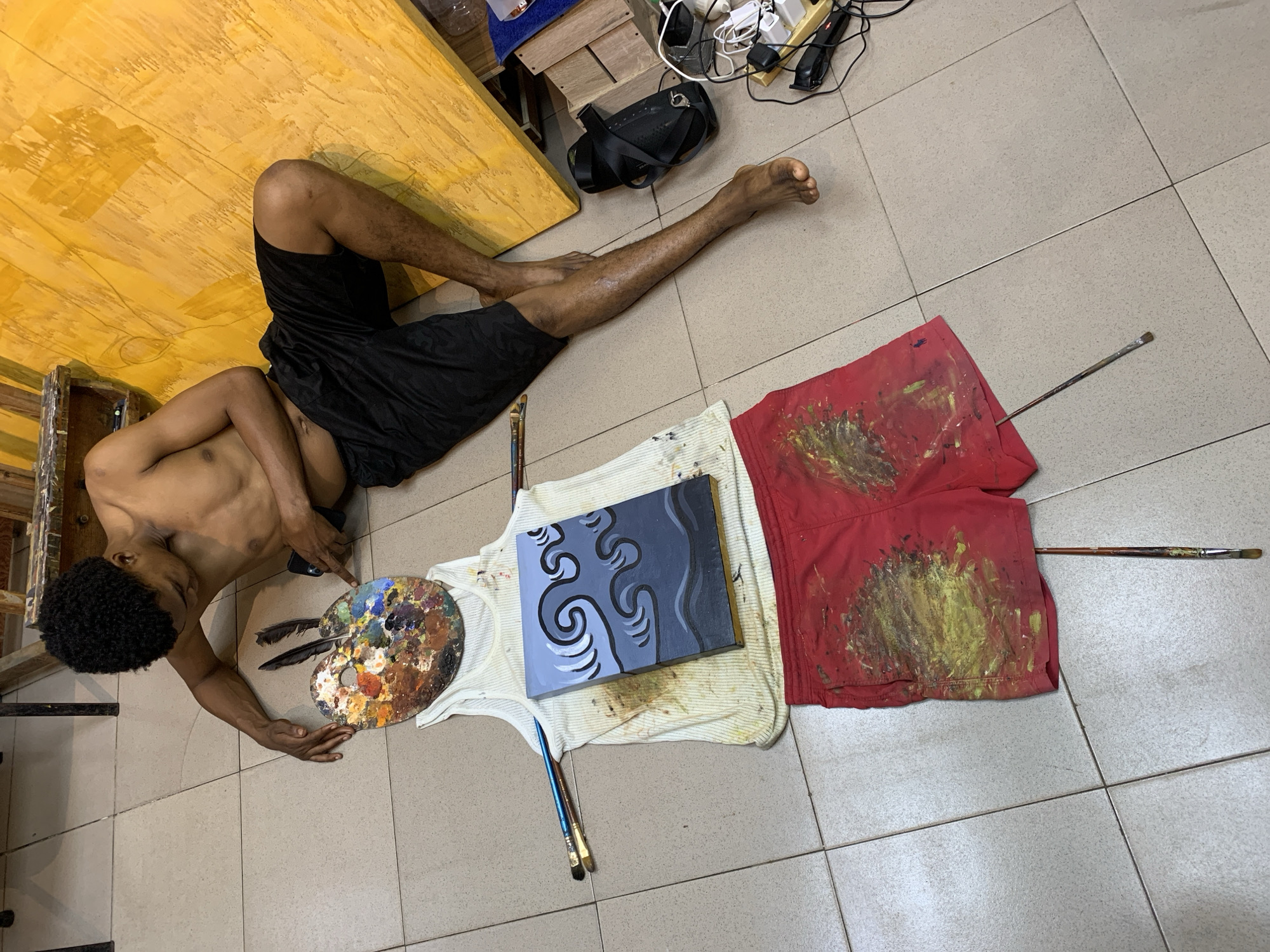 The image depicts an artist, shirtless and wearing black shorts, lying on a tiled floor in what appears to be an art studio. The artist is positioned next to a large, unfinished yellow canvas. A palette splattered with various colors of paint is near the artist's head, along with two feathers. In front of the artist, a white tank top and red shorts are arranged on the floor, creating a mannequin-like figure. A small painting with a wave motif is placed on the tank top. Several paintbrushes are scattered around the artist and the clothing. The environment shows evidence of artistic activity, with paint stains on the floor and clothing. Some electronic devices and a pair of sandals are visible in the background. The scene suggests a pause in the artistic process, with the artist resting amidst their work.
