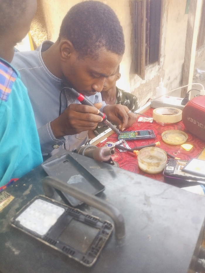An outdoor scene in Akwanga, Nigeria, depicts three individuals engaged in mobile phone repair. A dark-skinned male, positioned centrally, wears a light blue/grey long-sleeved shirt and focuses on soldering an open mobile phone's circuit board with a red-tipped soldering iron held in his right hand. His left hand stabilizes the phone's internal components. To his immediate left, a second individual's shoulder and back are visible, wearing a light blue shirt, partially observing the activity. Behind the primary repairman, a third dark-skinned male's face is visible, also observing.
The repair work occurs on a table covered with a red patterned cloth. Scattered across this table are various mobile phone components and repair tools: a disassembled phone with an exposed circuit board, another partially disassembled phone, several small screwdrivers with yellow handles, two clear plastic containers with yellowish residue (likely solder flux), a roll of beige masking tape, a red KOLEER brand portable speaker, a white multi-outlet power strip with an adapter, and a white plastic bottle. In the foreground, on a dark, metallic surface, a dismounted phone frame with a white rectangular component and a black phone case are visible alongside a curved metal bar. The background features a light-colored, rough-textured wall and a wooden window frame, suggesting an outdoor or semi-enclosed workshop environment under natural daylight.