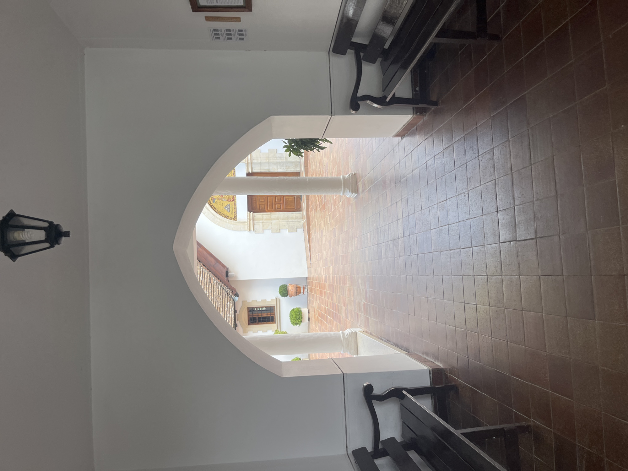 This is a photograph of an arched entryway leading to a courtyard. The entryway is covered, with white walls and a dark-tiled floor. On either side of the entryway are dark wooden benches. The courtyard beyond is paved with terracotta tiles. In the courtyard, there is a white building with a terracotta-tiled roof, a wooden door, and a window. There are also potted plants and a column visible. The photograph appears to have been taken during the daytime, with natural light illuminating the scene. The specific location in the city of Foini, Cyprus is not identifiable from the image alone, but the architectural style and outdoor setting suggest a Mediterranean or similar climate. There are no people visible in the image.