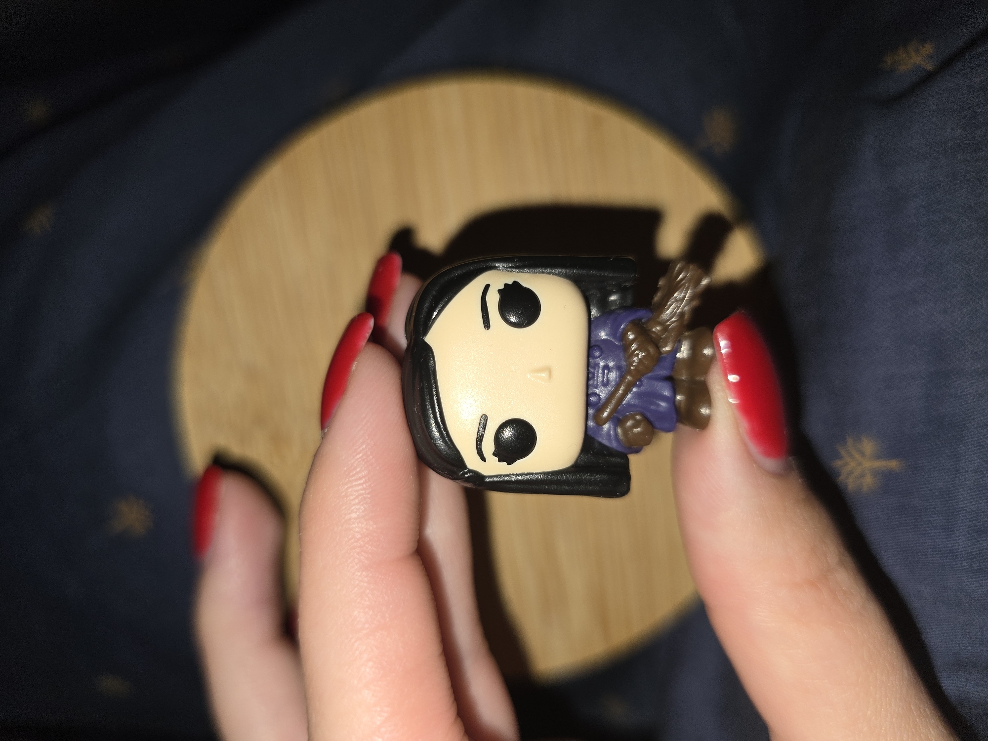 A close-up shot shows a miniature Funko Pop figure being held between the thumb and index finger of a person with bright red nail polish. The figure is a character with black hair, large black eyes, and a light-skinned face. It is wearing a dark purple robe and holding a brown broomstick in its hands. The figure is positioned over a light brown, circular surface, which is itself resting on a dark blue, textured fabric background. The background fabric has subtle, repeating gold patterns. The image is taken from a high angle. There is no identifiable location in Petropavlivska Borshchahivka, Ukraine present in the image.