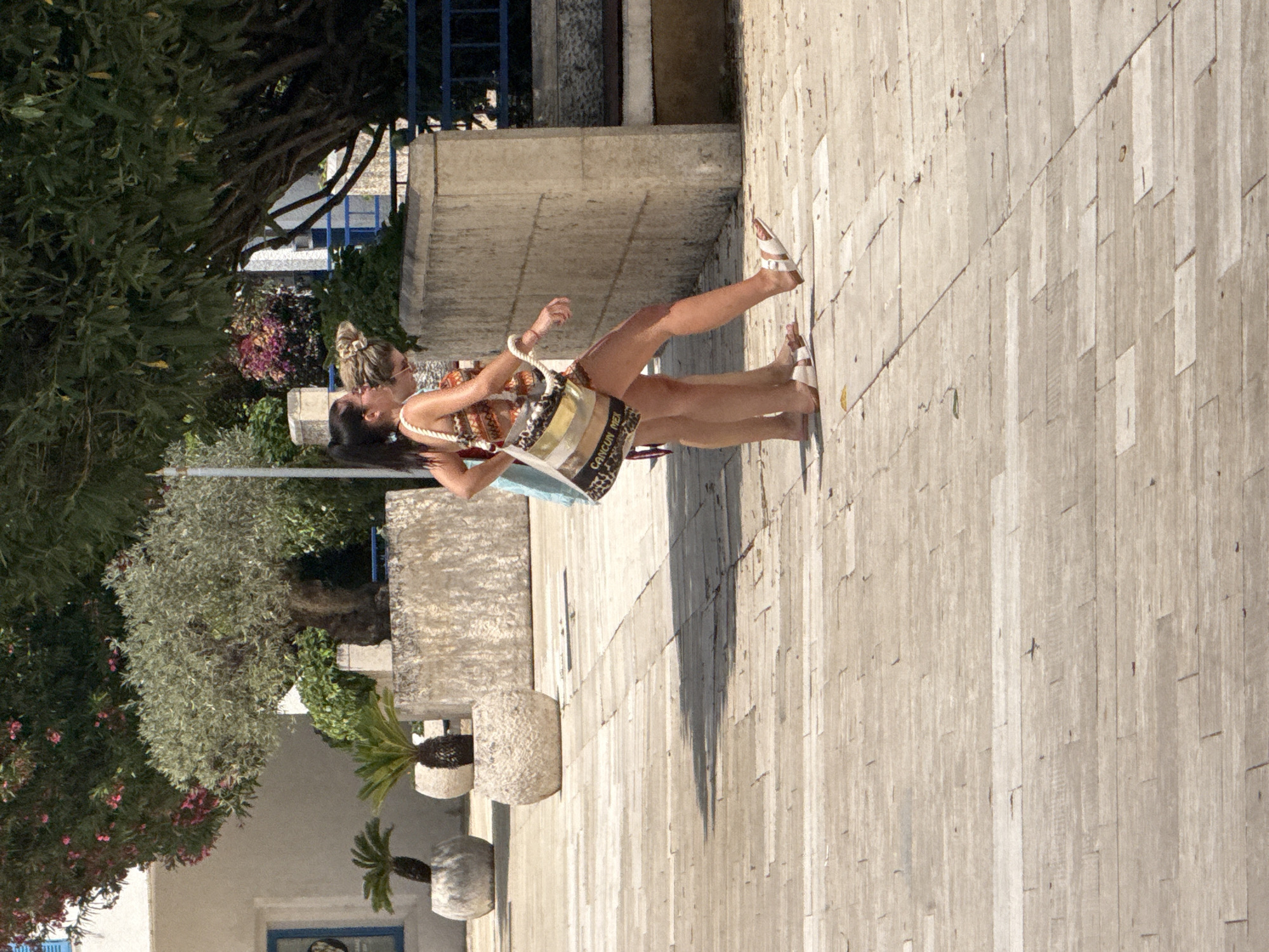 Two women are walking on a paved courtyard in Boreti, Montenegro. The woman in the foreground, wearing a patterned dress and white sandals, carries a woven bag with the text "CANCUN MEX" visible. The woman behind her, wearing a light blue dress and flip-flops, is partially obscured. They are walking towards the right of the frame. In the background, there are potted plants, trees, and stone structures, with a building with blue shutters visible to the left. The ground is paved with light-colored rectangular stones.