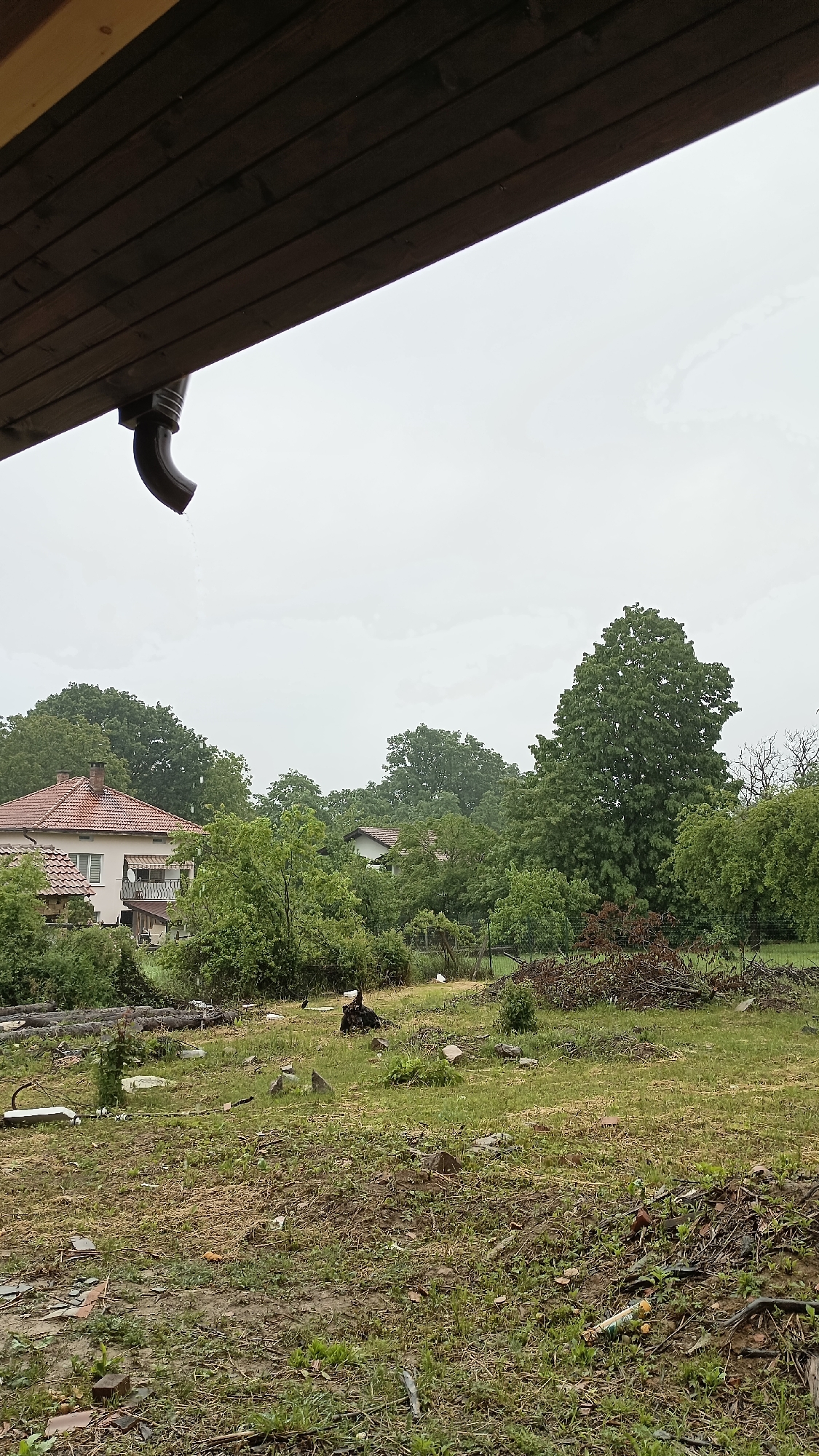 The image, taken in Mihaylovtsi, Bulgaria, shows a view from under a wooden roof overhang with a gutter discharging rainwater. In the background are several houses partially obscured by trees. The foreground is a yard with grass and scattered debris, including pieces of wood and other materials. The sky is overcast. There are no people visible in the frame.