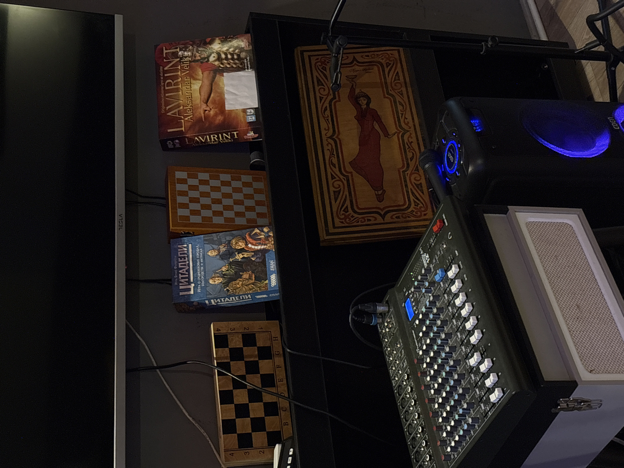 The image shows a cluttered shelf with various objects. There is a black Tesla monitor on the left side. On the shelf are a board game called "LAVIRINT Aleksandar Veliki", two chess boards, a board game called "ЦИТАДЕЛИ", an ornately framed picture, an audio mixer, and a speaker with blue lights. There is no information to suggest that the photo was taken in Kragujevac, Serbia. The image shows objects on a shelf, and no people or actions are visible.
