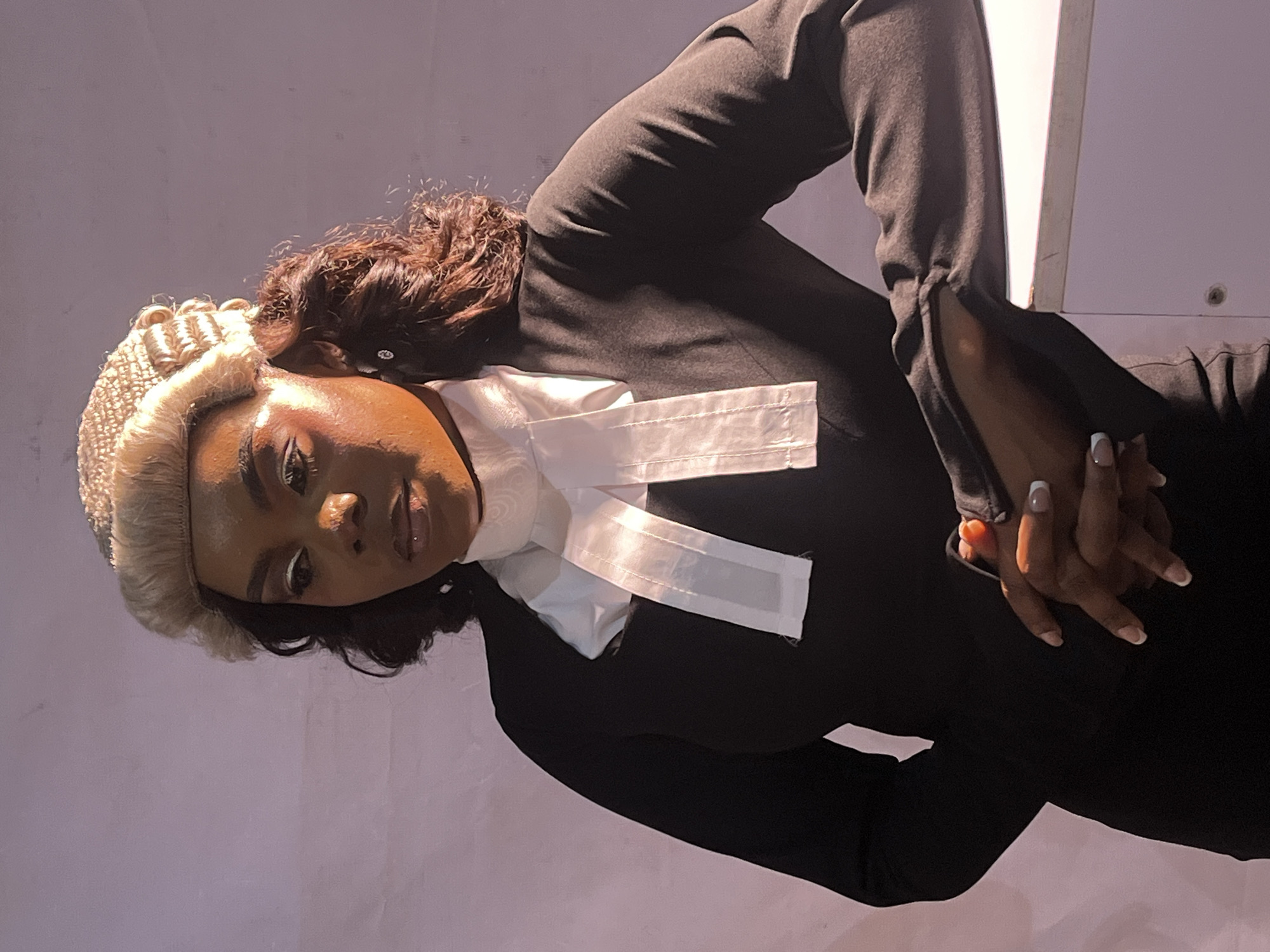 A woman is photographed from the chest up, posed with her arms crossed and resting on a white surface. She is wearing a black robe, a white bib with hanging bands, and a barrister's wig. Her head is slightly tilted, and she is looking downward and to her left. Her makeup is visible, with shimmering eyeshadow and lip gloss. The background is a plain, muted purple and pink gradient. The lighting casts a warm glow on her face and shoulders, highlighting the texture of the wig and the sheen of her clothing. The image appears to be taken in a studio setting, possibly in Jos, Nigeria, given the implied location.
