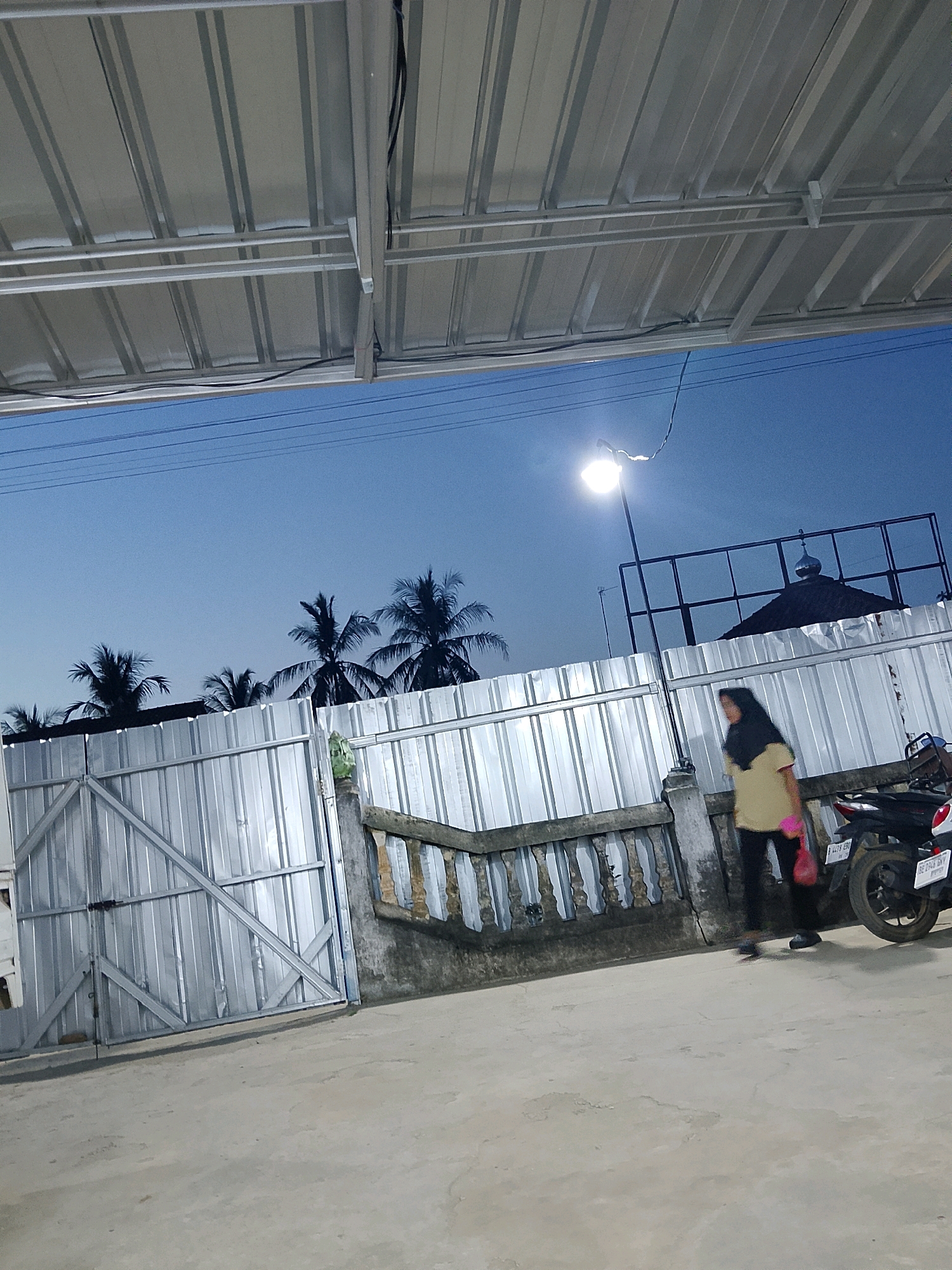 In Negeri Agung, Indonesia, at dusk, a woman in a yellow shirt and black pants walks on a concrete path in front of a corrugated metal fence. A motorcycle is parked to her right. Above the fence, palm trees and a mosque dome are visible against the twilight sky, illuminated by a streetlamp. A metallic roof structure extends over the scene.