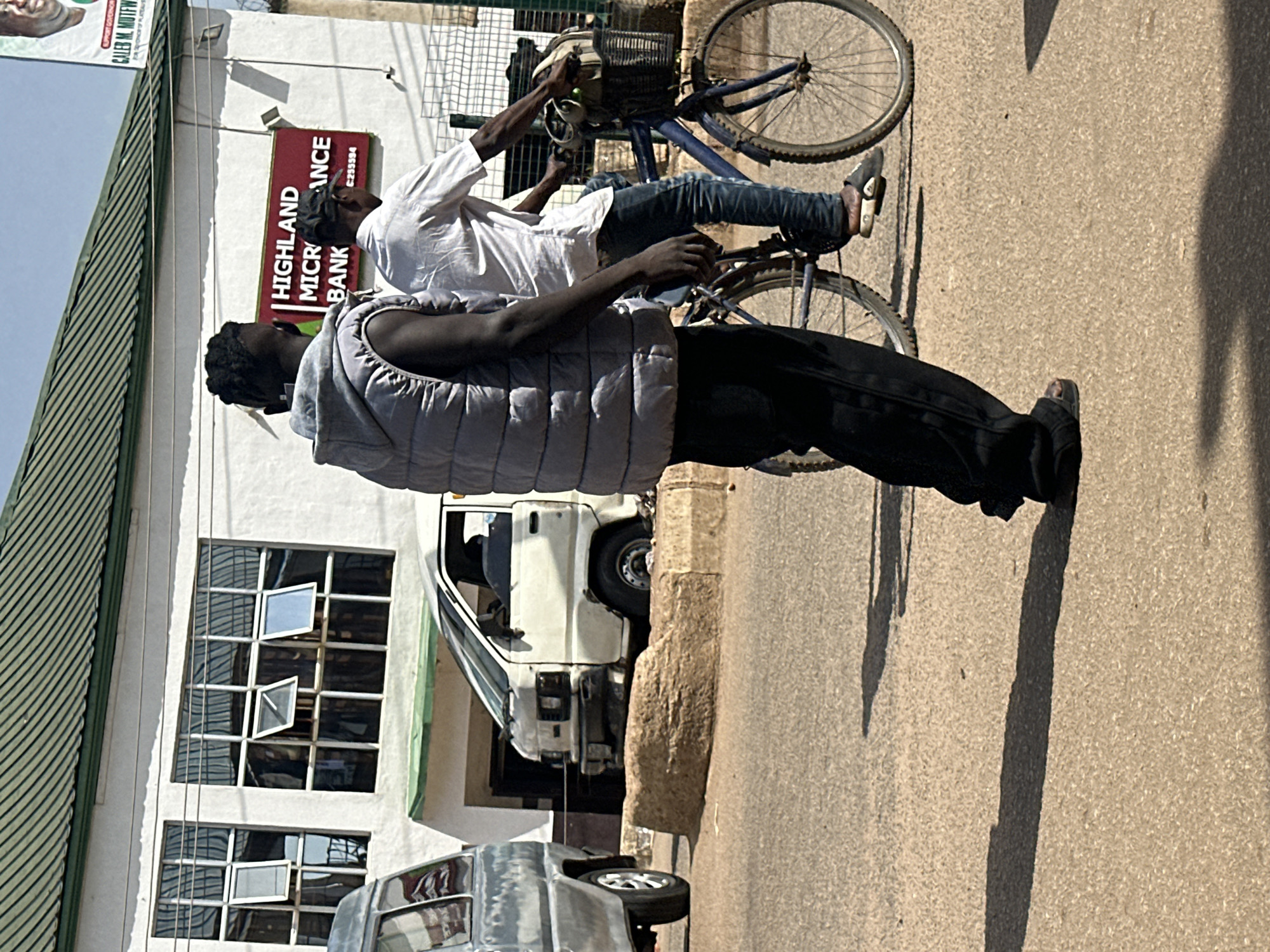A medium shot captures two men walking in the city of Bukuru, Nigeria. The man in the foreground, viewed from behind, wears a grey puffer vest over black pants and sandals. He is walking on a paved road with a shadow cast on the ground. To his right, another man is riding a blue bicycle, also casting a shadow. He wears a white short-sleeved shirt, blue jeans, and sandals. The bicycle has a basket on the handlebars and a bag is placed inside it. In the background, a white building with a green roof and large windows is visible. A sign on the building reads "HIGHLAND MICROFINANCE BANK." A white van is parked in front of the building, and another vehicle, partially visible on the left, is also present. The scene appears to be a street in a commercial area, under bright daylight conditions.