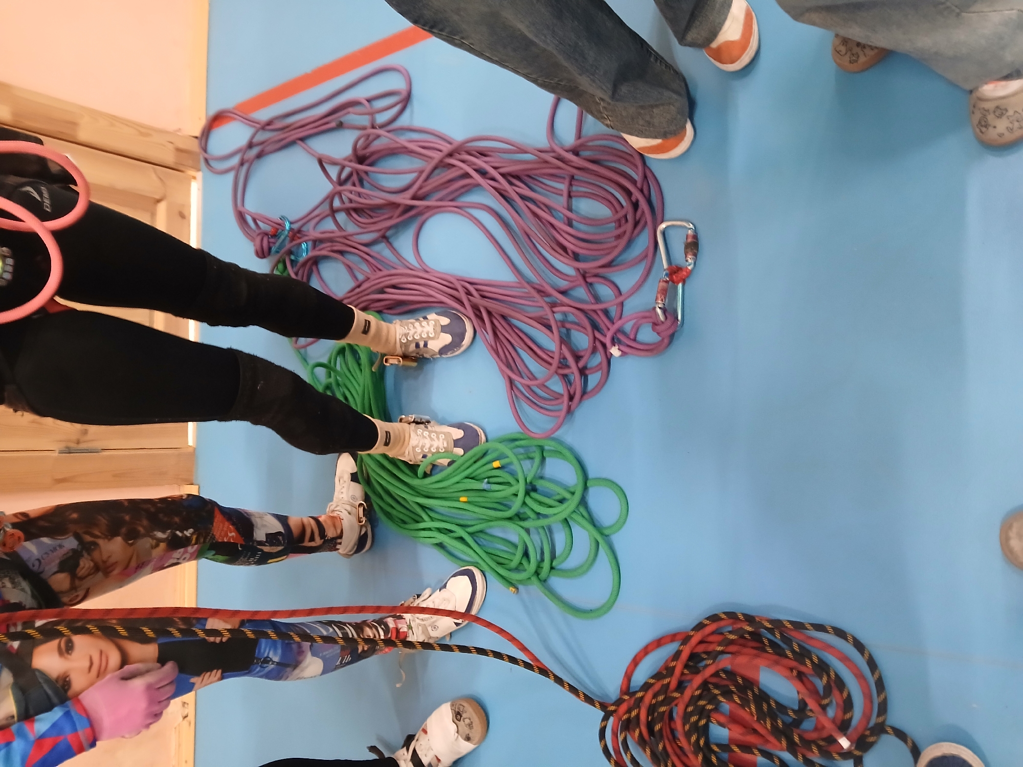 The image, taken from an elevated perspective, shows a group of people standing on a blue floor. Several coiled ropes of varying colors (red, green, and purple) are scattered on the floor. Some of the people are wearing athletic shoes and leggings; one pair of leggings features a collage of faces. A climbing harness is visible on one person. The precise location within Berezniki, Russia, is not discernible from the visual information provided.