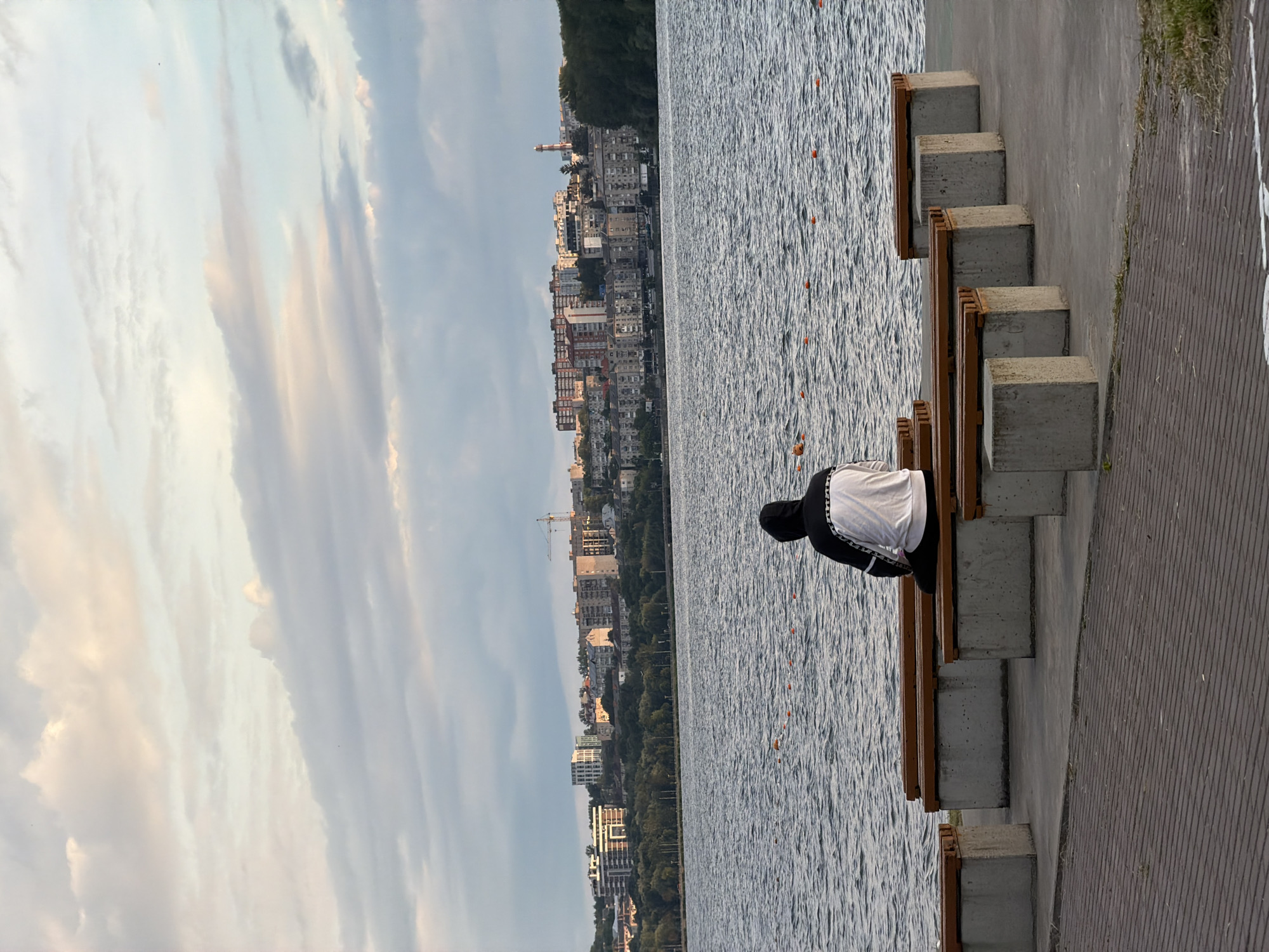 A person wearing a black hooded sweatshirt with white and black accents sits on a wooden bench facing a lake in Ternopil, Ukraine. The lake has ripples on its surface and is dotted with orange buoys. In the background, across the lake, is a cityscape with various buildings, trees, and a construction crane. The sky is cloudy with soft lighting. The foreground consists of a paved walkway and several more benches, some empty and some with concrete bases. The scene appears to be an outdoor public space.