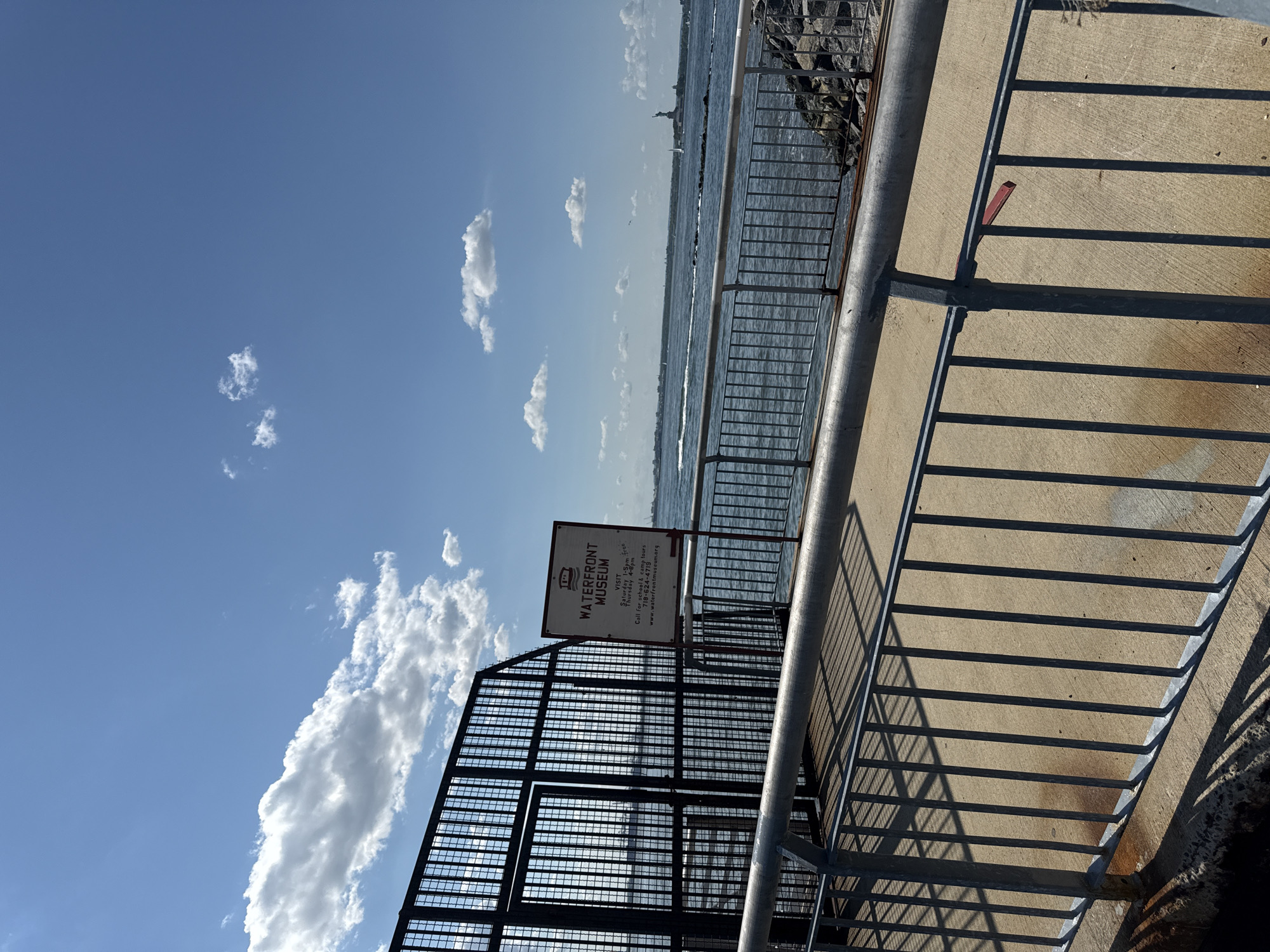 This is an eye-level, wide shot of a paved walkway along a waterfront, with a metal railing in the foreground. The walkway leads to a large, black metal fence with a gate. To the right of the fence, a sign reads "WATERFRONT MUSEUM" and provides visiting hours, a phone number, and a website. Beyond the walkway and fence, a body of water is visible, with a distant shoreline and the Statue of Liberty on an island. Several sailboats and ships are scattered across the water. The sky above is a clear blue with scattered white clouds. The scene appears to be in the vicinity of the Statue of Liberty in New York Harbor.