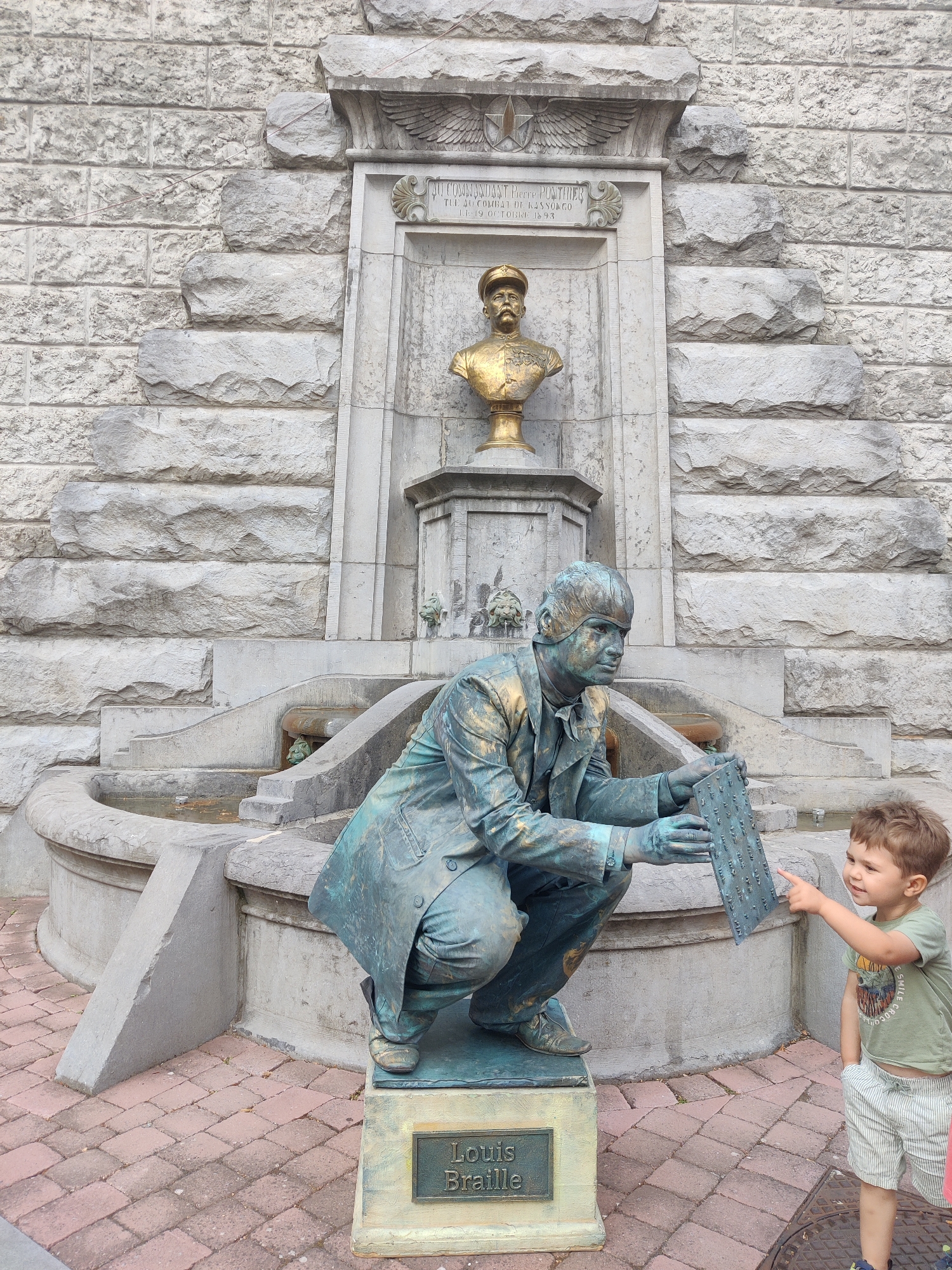 The image depicts a street scene in front of a stone building, likely in Marche-en-Famenne, Belgium, featuring a living statue and a young child. The living statue, painted in a metallic green and gold patina, is posed in a crouched position, holding a Braille slate with raised dots. It is standing on a pedestal with a plaque that reads "Louis Braille." To the right of the statue, a young boy in a green t-shirt and striped shorts is pointing at the Braille slate, seemingly interacting with the statue. Behind the statue, a tiered stone fountain structure is visible, integrated into the building's facade. The building itself is constructed from large, roughly cut stone blocks. The ground is paved with red and grey brickwork. The lighting suggests it is daytime.