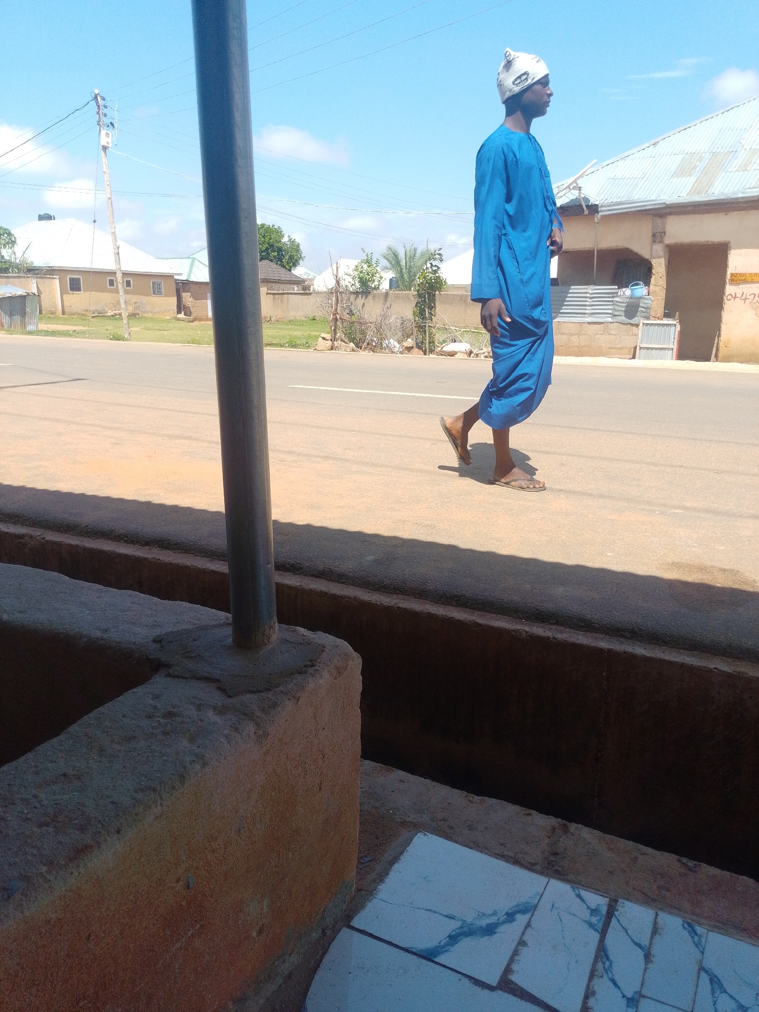 A person wearing a blue robe and a white patterned hat walks on the right side of a road. The person is facing to the left and is wearing sandals. The road is paved and has a white line marking the lane. In the background, there are buildings and trees. The sky is blue with some clouds. In the foreground, there is a concrete structure and a metal pole.