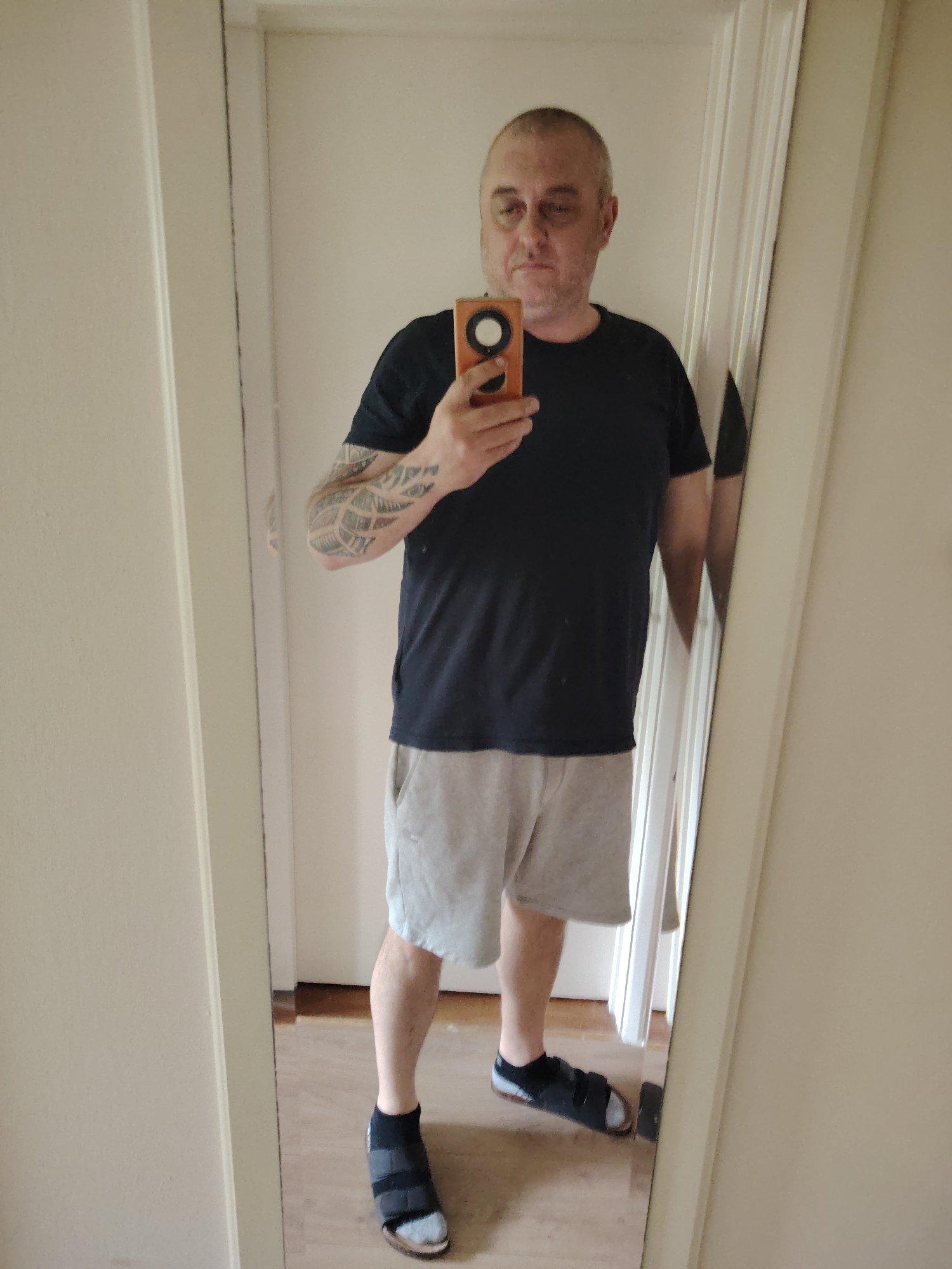 A middle-aged male, appearing to be in his 40s to 50s, is depicted in a mirror selfie taken indoors in Huntingdonshire, United Kingdom. He has short, receding grey hair and facial stubble. He is wearing a plain black short-sleeved t-shirt and light grey shorts. A large, abstract, possibly tribal-style tattoo is visible on his left forearm. He holds an orange-cased smartphone in his right hand, pointing it towards the mirror to capture his reflection. On his right foot, he wears a black sock and a black sandal with two broad straps. On his left foot, he wears a light grey sock and a black orthopedic-style post-operative boot or sandal with multiple velcro straps. The background shows light-colored walls and a light wooden or laminate floor, with a white door frame visible to the left and reflected on the right.