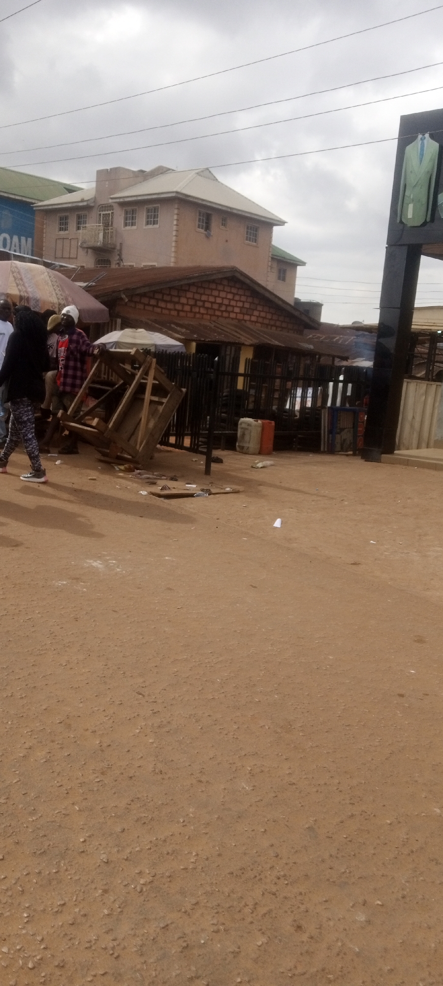 The image shows a street scene in Sabon Tasha, Nigeria. The foreground consists of a dirt road with scattered debris. In the midground, there are several people, including one wearing a plaid shirt and another with patterned pants. A wooden structure appears to be overturned near a metal fence. Behind the fence are small buildings with metal roofs and a brick building with a pitched roof. A tall building is visible in the background. A large sign featuring a suit is on the right. The sky is overcast.